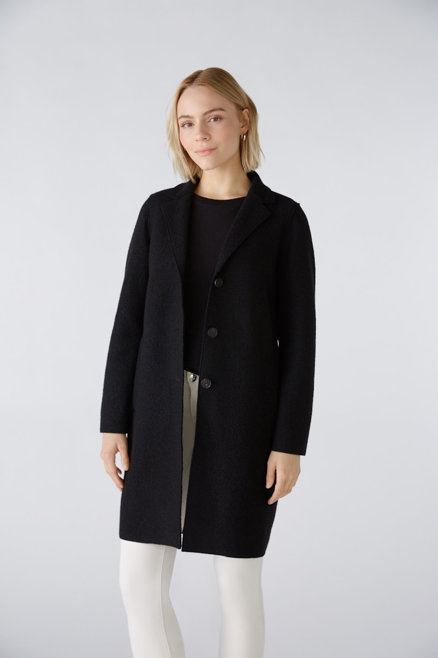 Gezeigt wird a woman wearing a black coat. The coat features a button closure and a tailored fit, perfect for a stylish look. It is paired with a simple top and light-colored pants, highlighting the coat as the main focus.