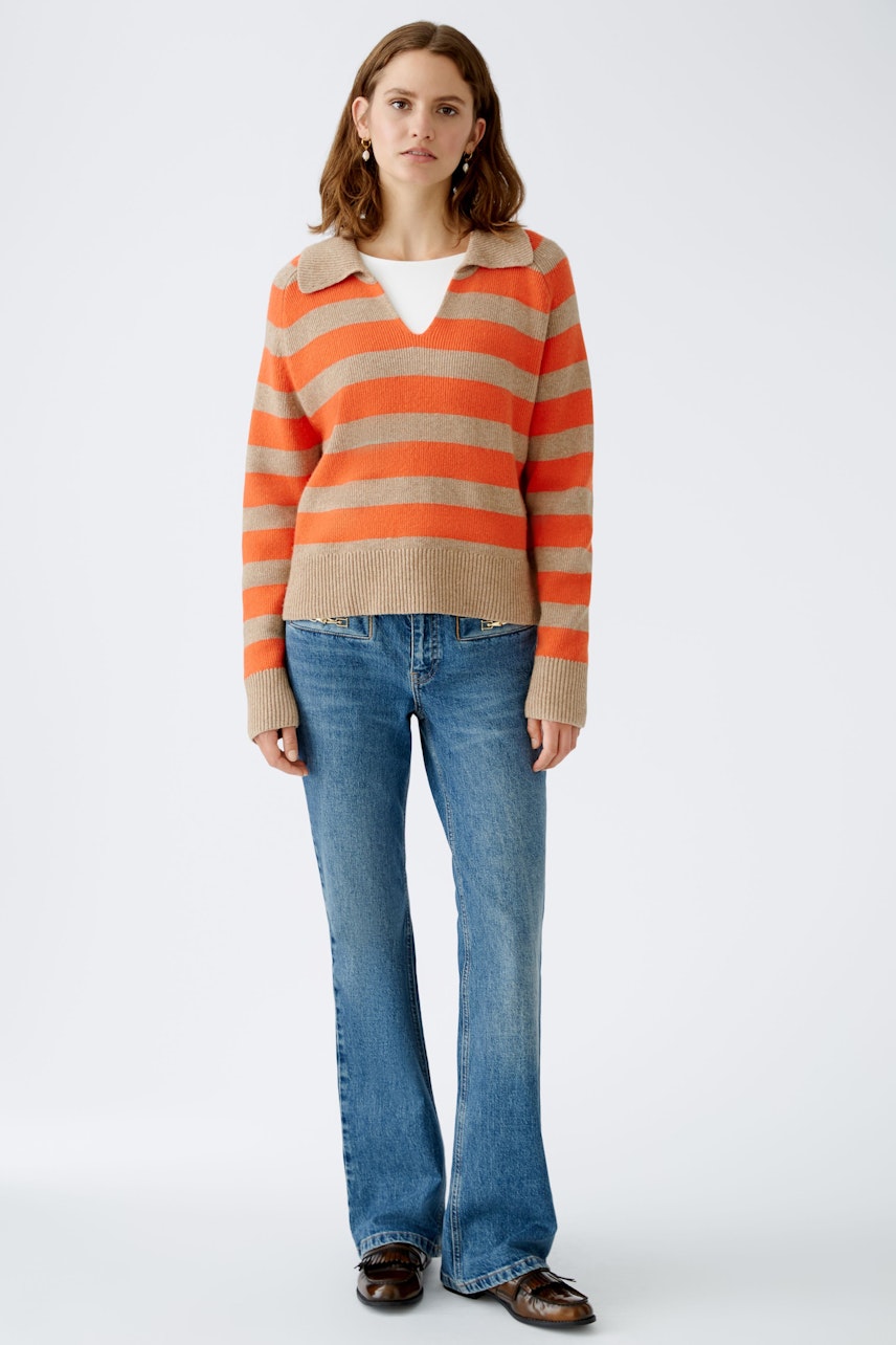 This image features a woman wearing a stylish outfit that includes a vibrant striped sweater in shades of orange and beige, complemented by a classic white top underneath. She pairs this with flared blue jeans, creating a trendy and casual look. The outfit is perfect for a relaxed day out or a cozy gathering, showcasing a blend of comfort and fashion.