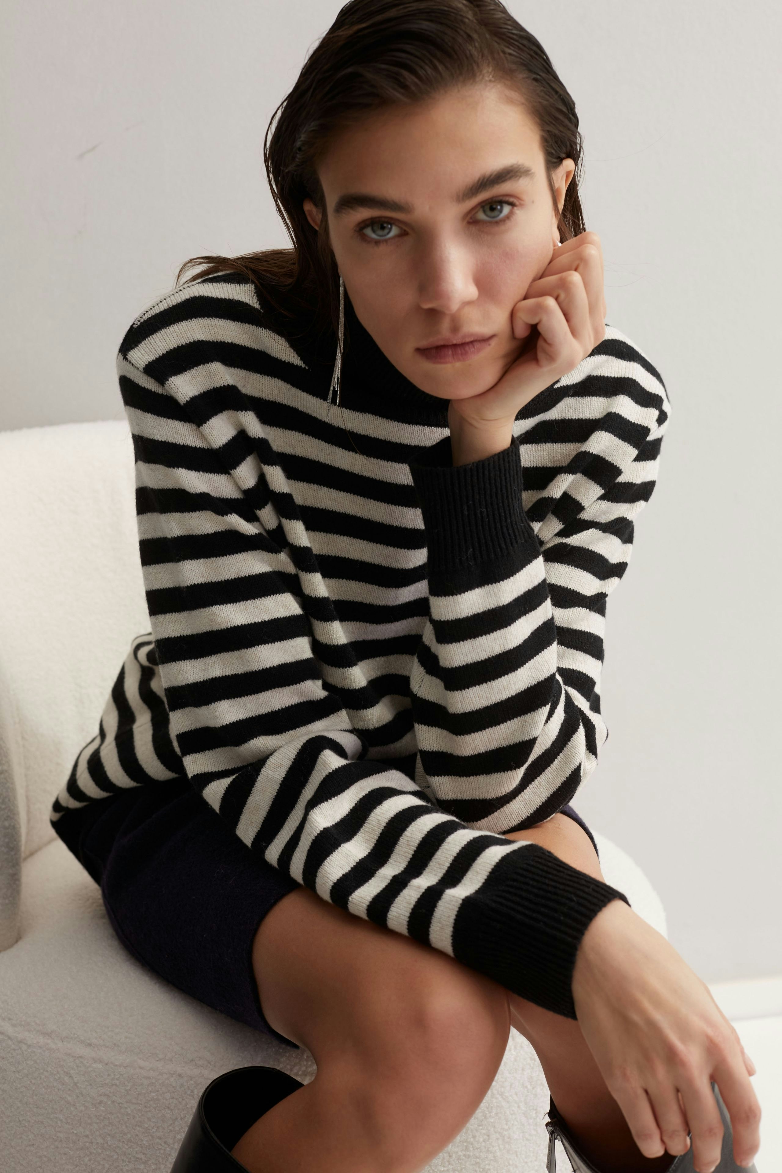 Damen-Pullover: Frau in gestreiftem Pullover mit schwarzem Bund sitzt nachdenklich auf einem Hocker.