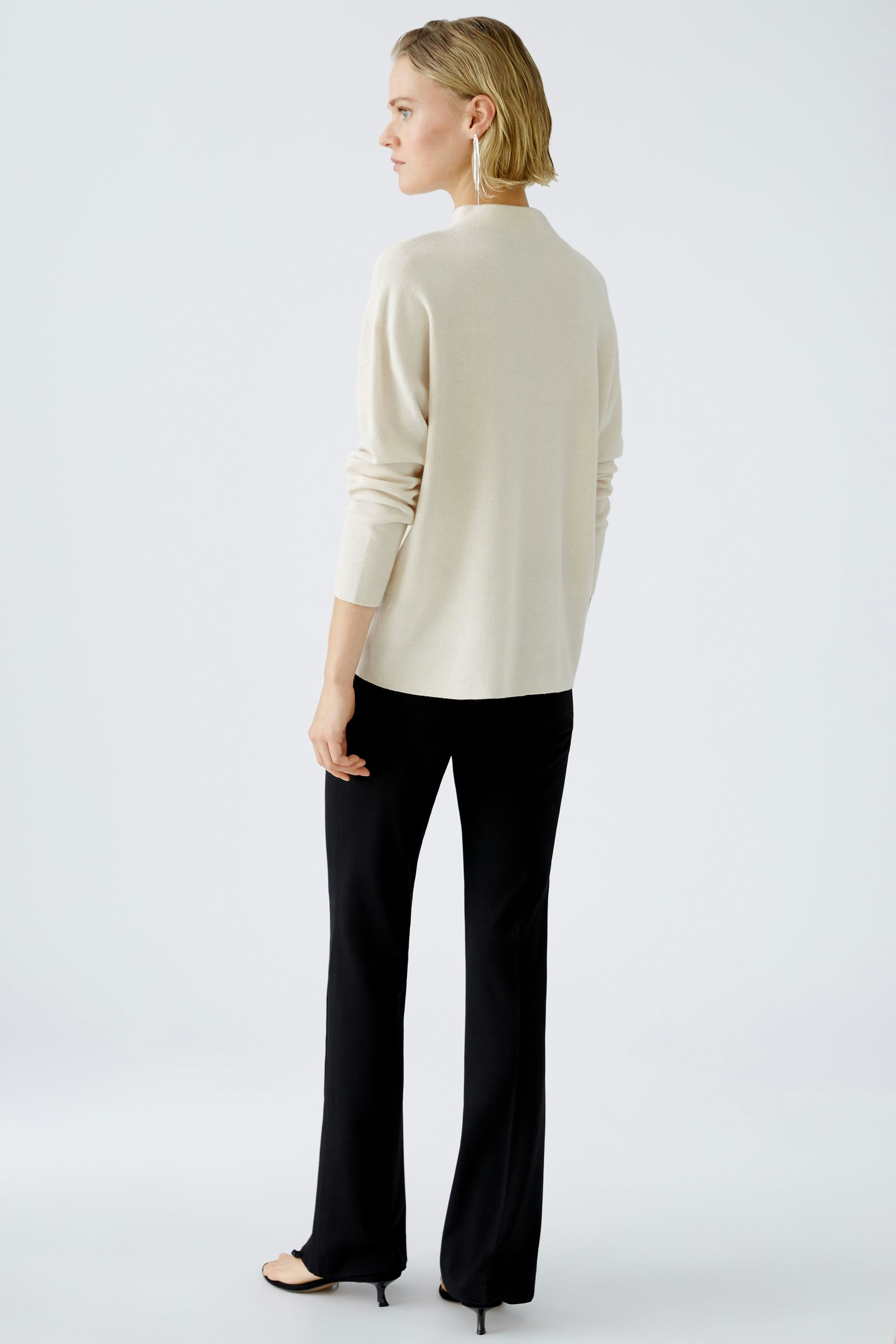 Modern women's pullover: A woman in a cream pullover and black pants stands confidently.