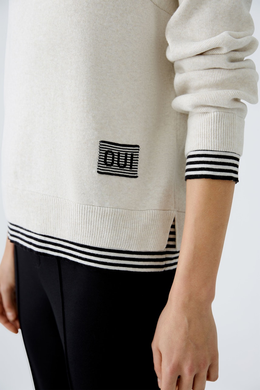 Stylish women's pullover: A woman in a cream pullover with striped cuffs and a logo detail.