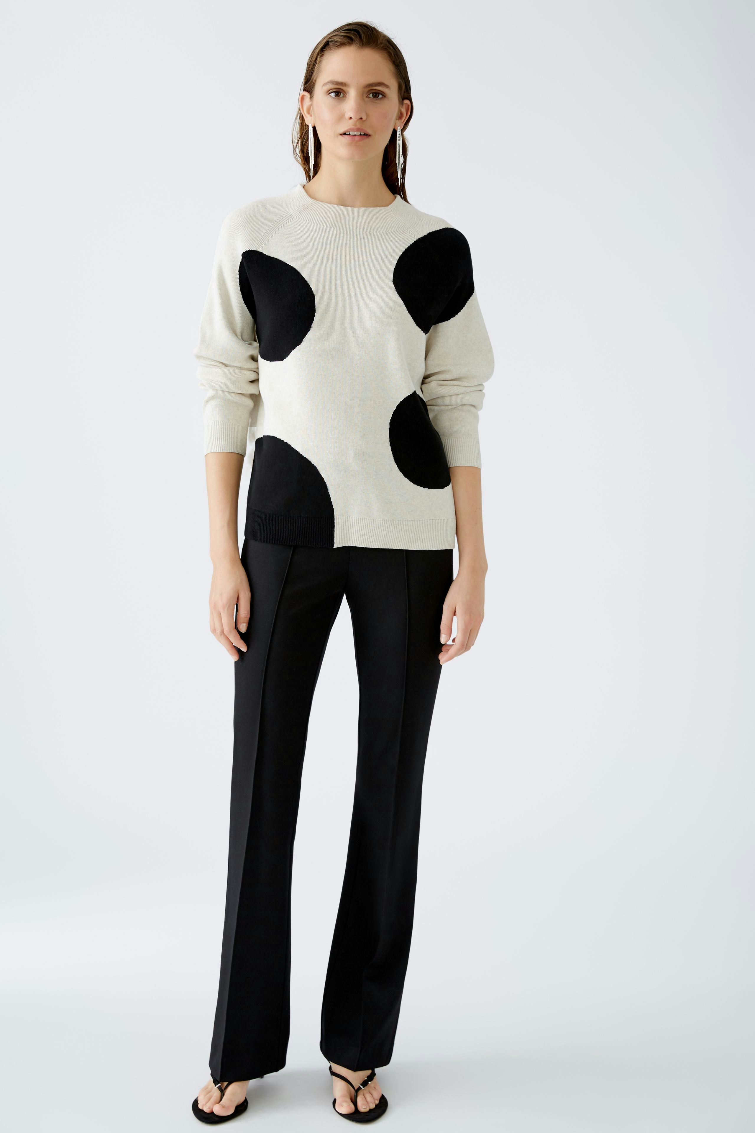 Modern pullover: Woman in a light pullover with black dots and stylish black pants stands confidently.