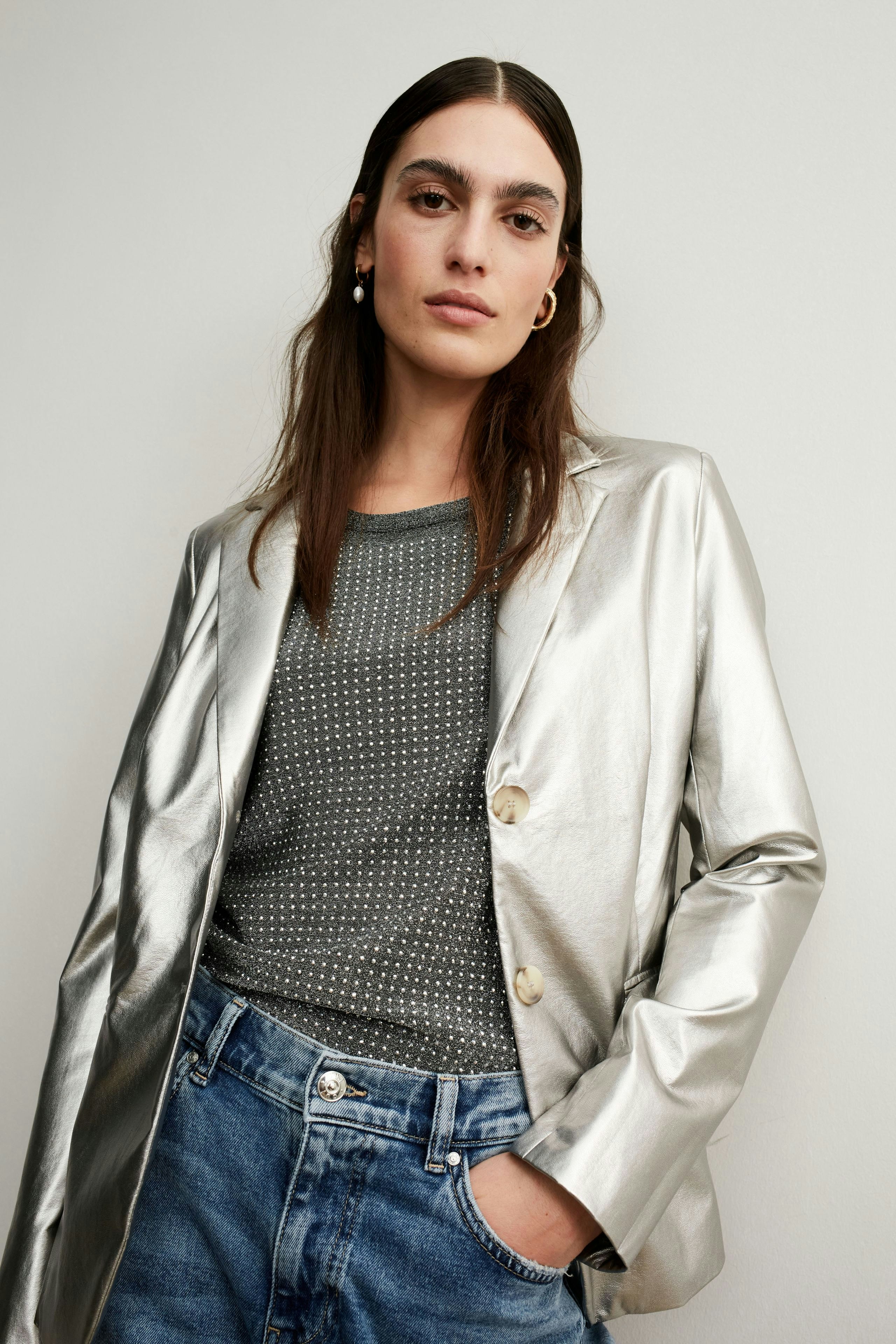 Stylish women's blazer: A woman in a shiny silver blazer over a dotted pullover, paired with casual jeans.