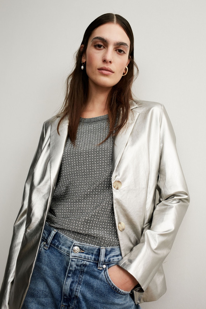 Stylish women's blazer: A woman in a shiny silver blazer over a dotted pullover, paired with casual jeans.