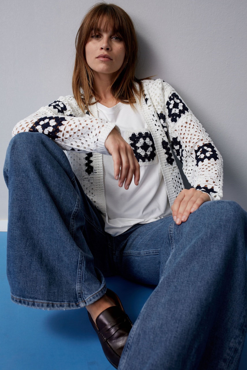 Stylish women's cardigan: A woman in a white and navy crochet cardigan sits confidently in wide-leg jeans.