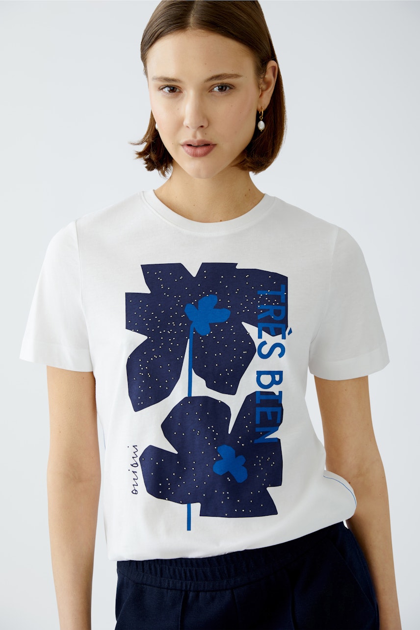 Damen-T-Shirt: Junge Frau trägt ein weißes T-Shirt mit blauen Blumenmotiven und lässigem Schnitt.