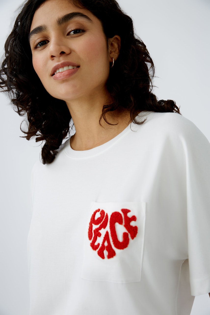 Stylish women's T-shirt: A woman in a white tee with a red 'PEACE' pocket design, radiating positivity.