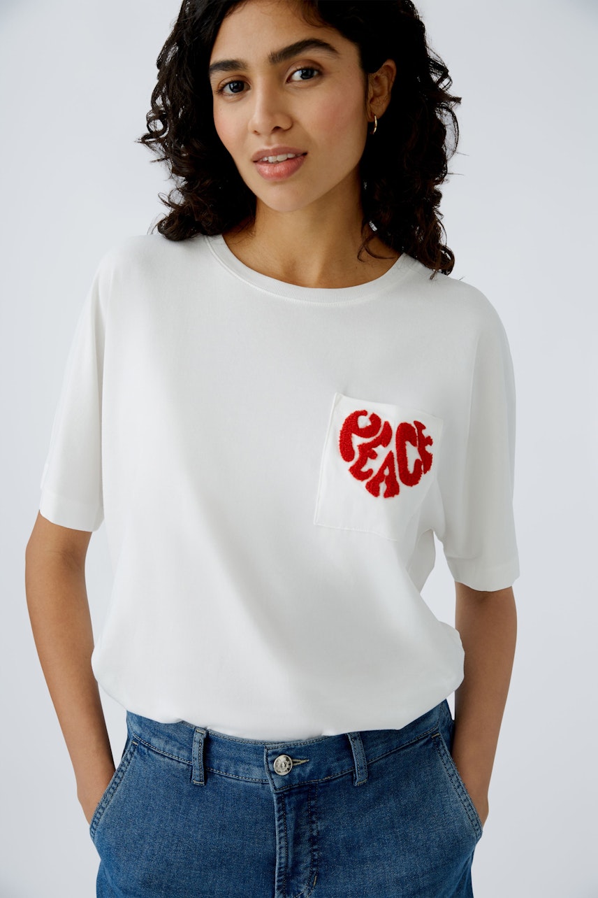 Stylish women's T-shirt: A woman in a white tee with a red 'PEACE' heart pocket design.