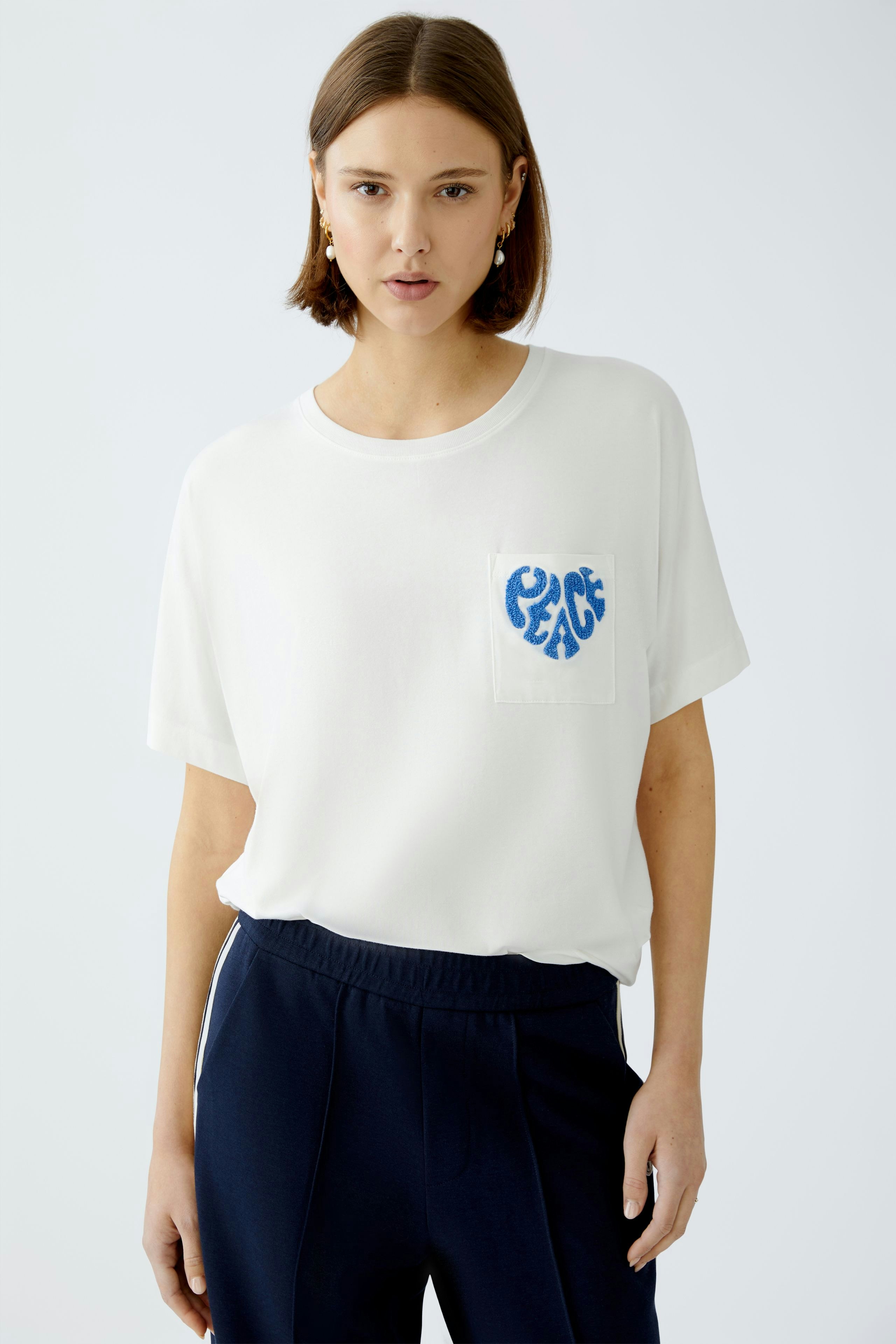 Bequemes Damen-T-Shirt: Frau trägt ein weißes T-Shirt mit blauer "PEACE"-Stickerei auf der Brust.