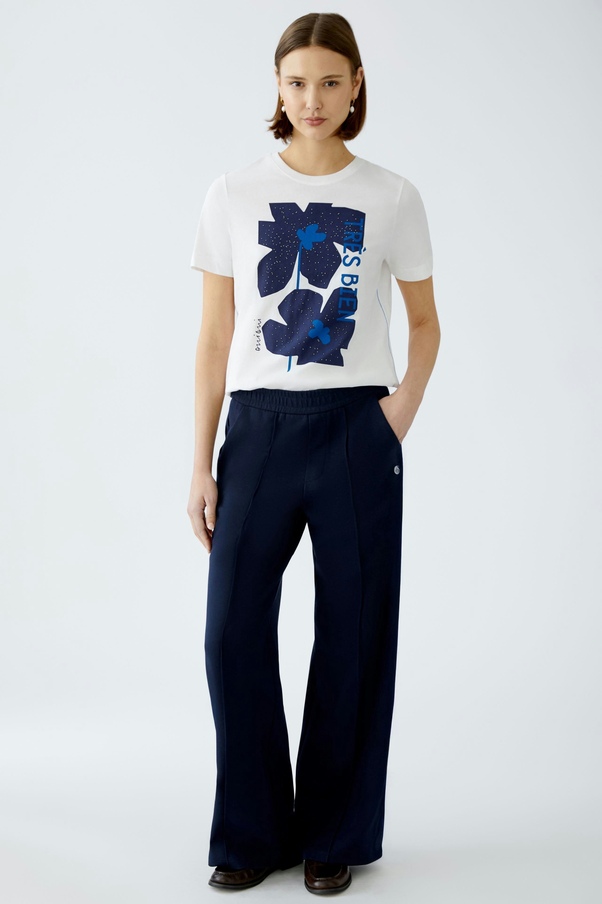 Damen-T-Shirt: Junge Frau trägt ein weißes T-Shirt mit blauem Grafikdruck und lässigen, weiten Hosen.