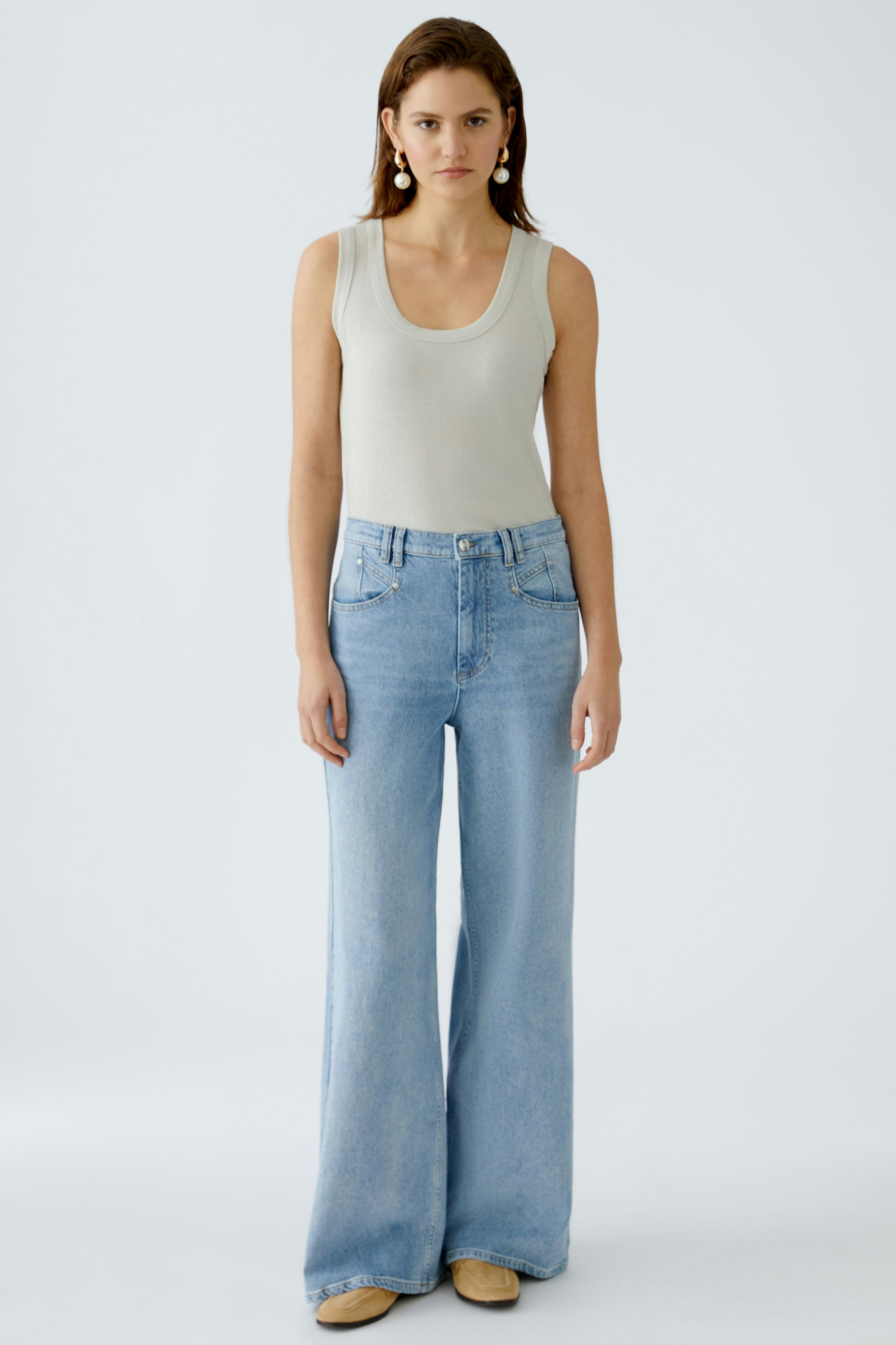 Stylish women's top: A woman in a light tank top paired with wide-leg jeans stands confidently.