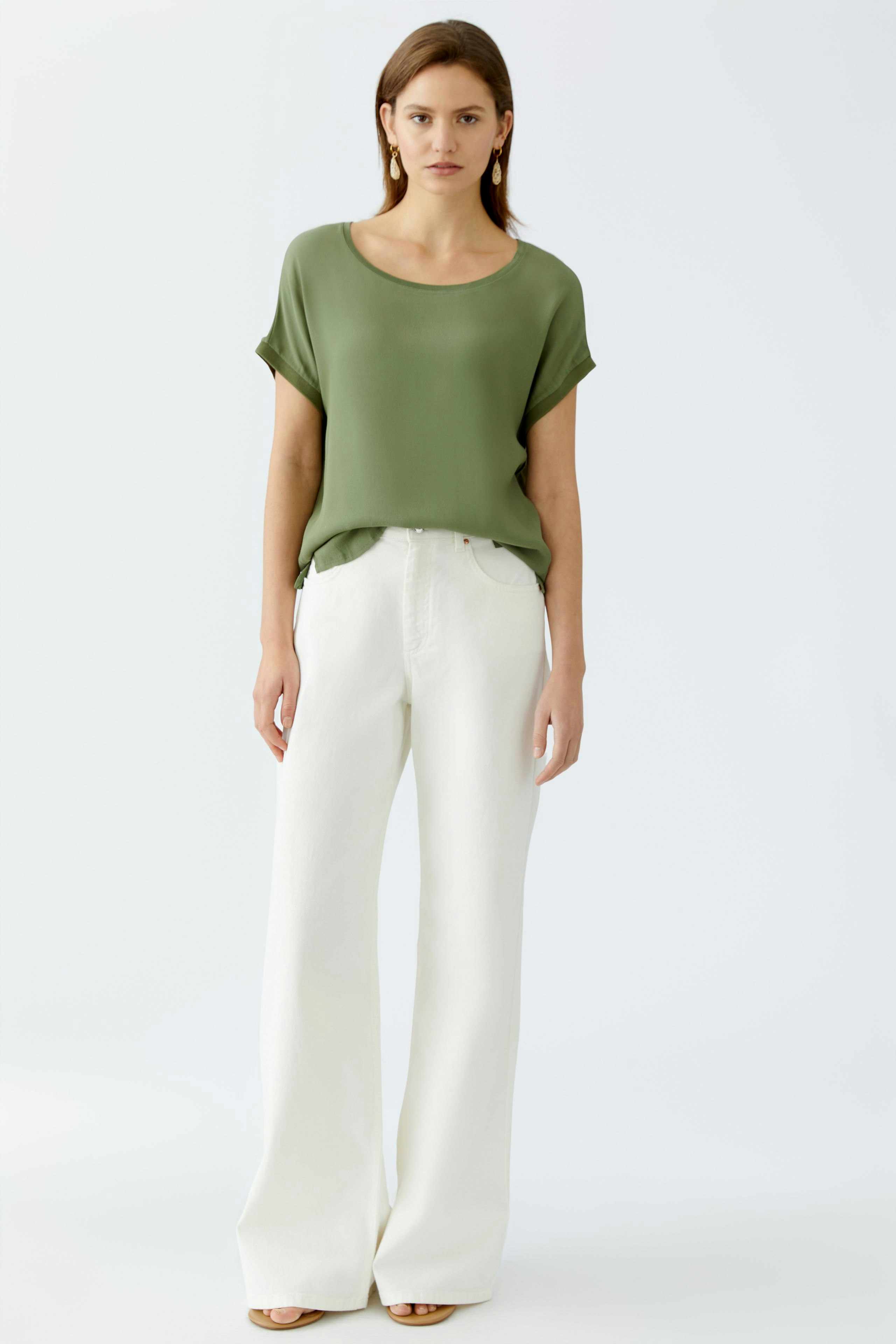 Luftiges Blusenshirt: Junge Frau in grünem Shirt und weißen, weiten Hosen steht entspannt.