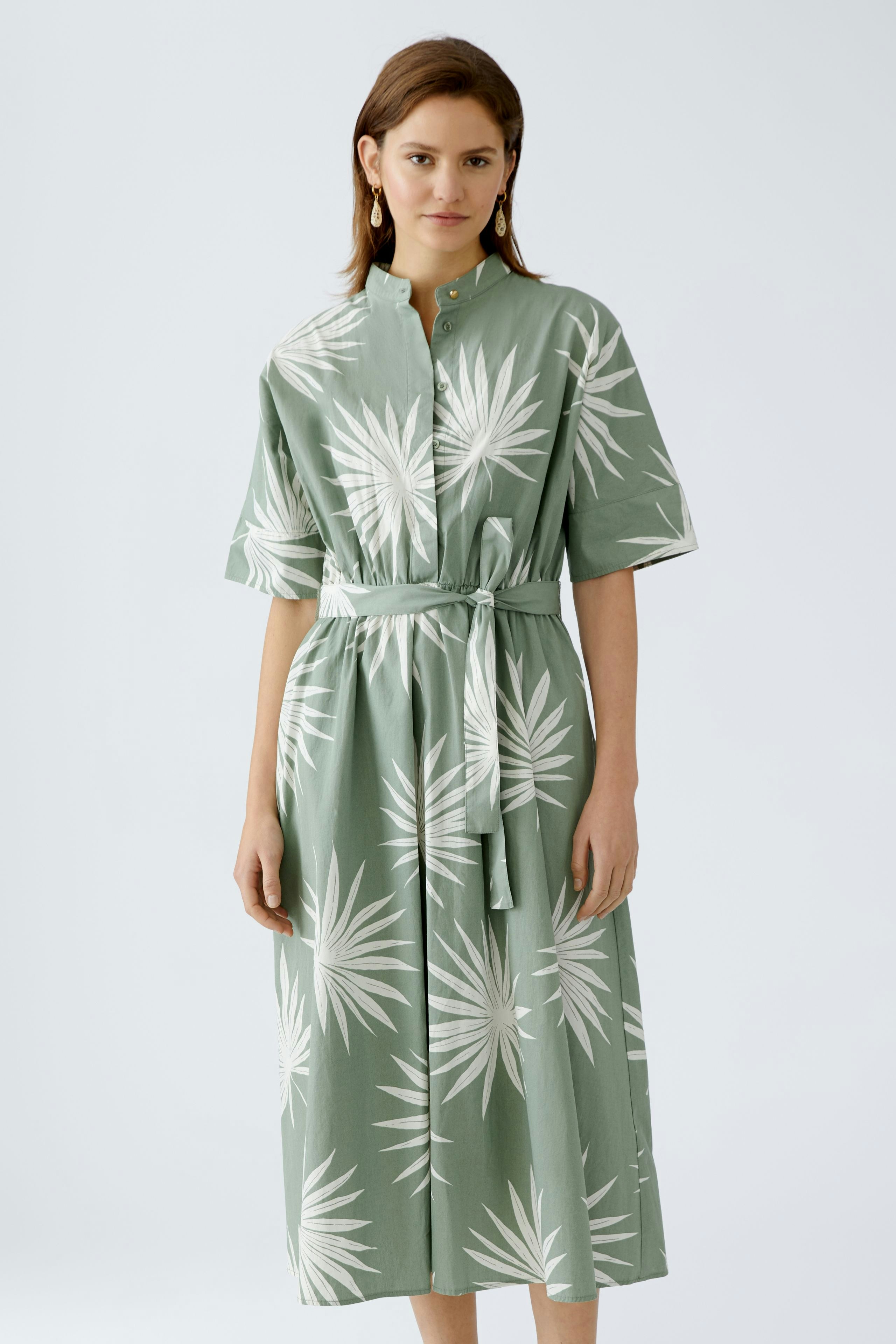Elegantes Midikleid: Frau in grünem Kleid mit Palmenmuster und Taillengürtel steht selbstbewusst.