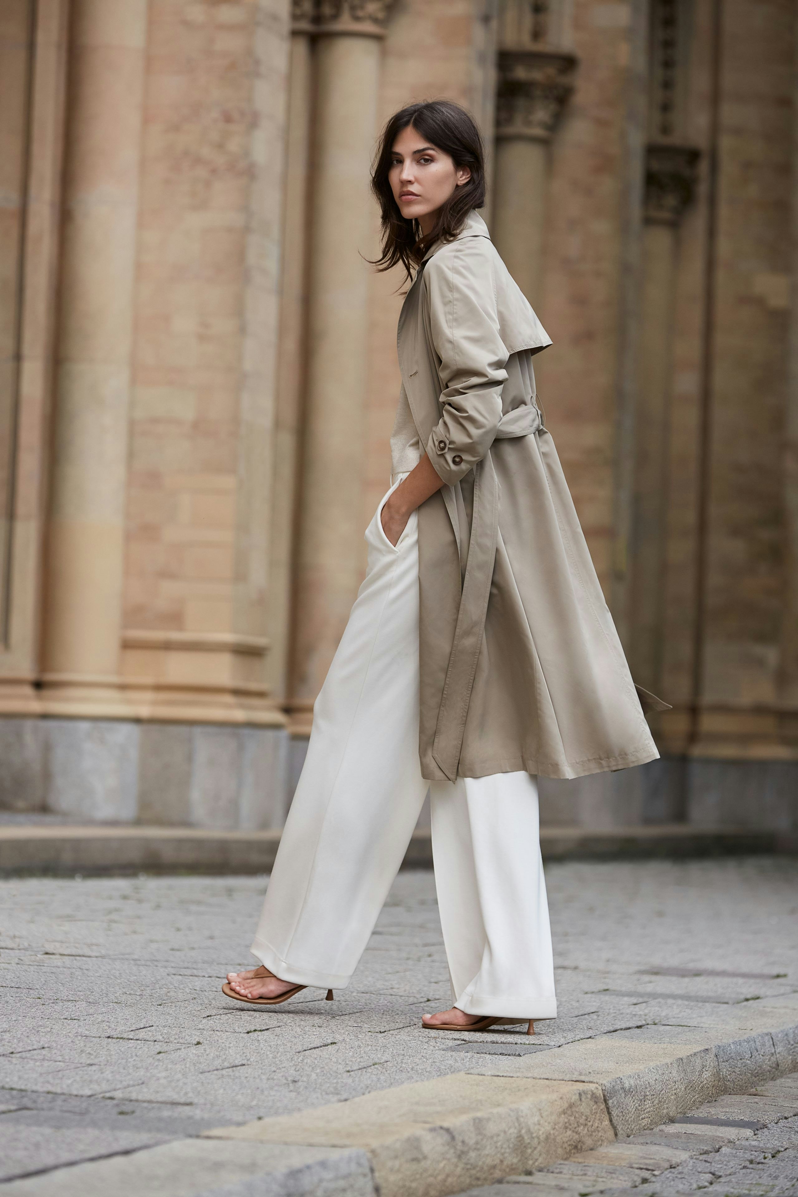 Stylish trench coat: A woman in a beige trench coat paired with white trousers stands confidently.