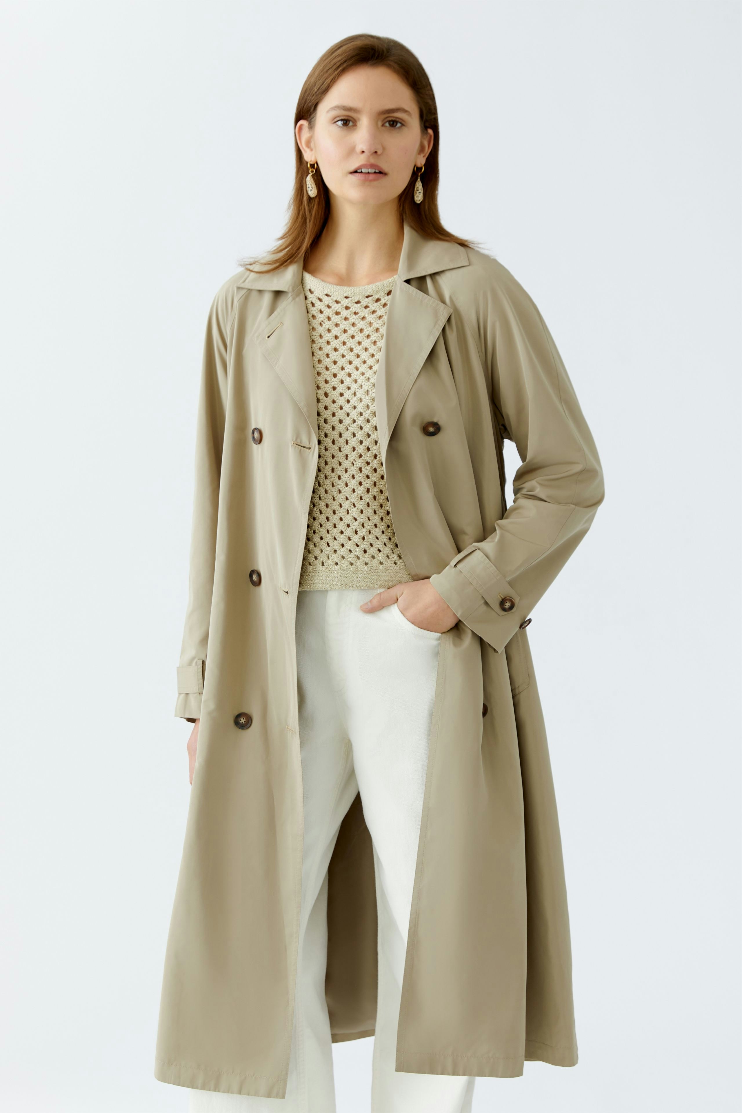 Stylish trench coat: Confident woman in a beige trench coat over a polka dot pullover and white pants.