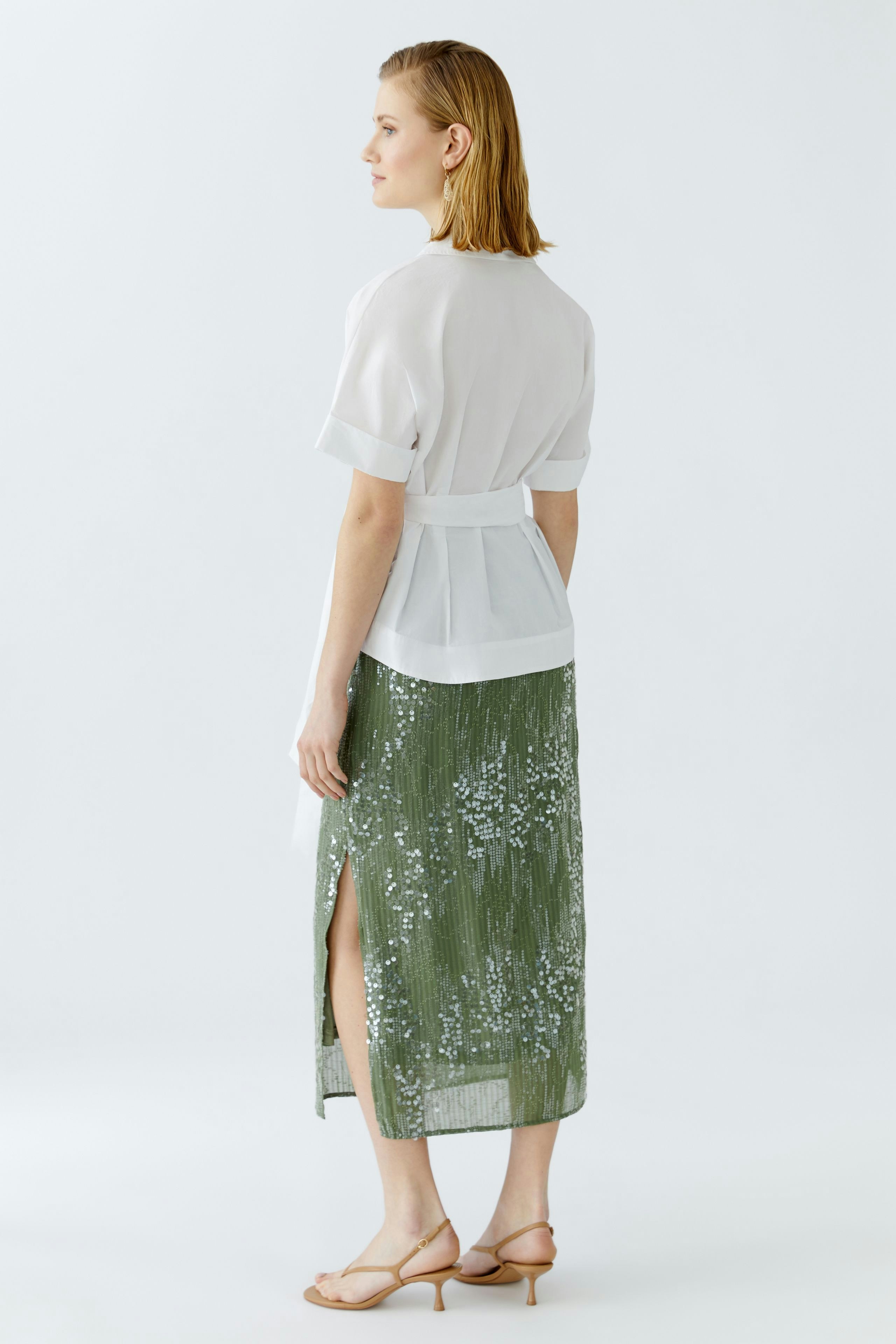 Elegante Damen-Bluse: Frau in weißer Bluse mit Gürtel und glitzerndem Rock, von hinten betrachtet.