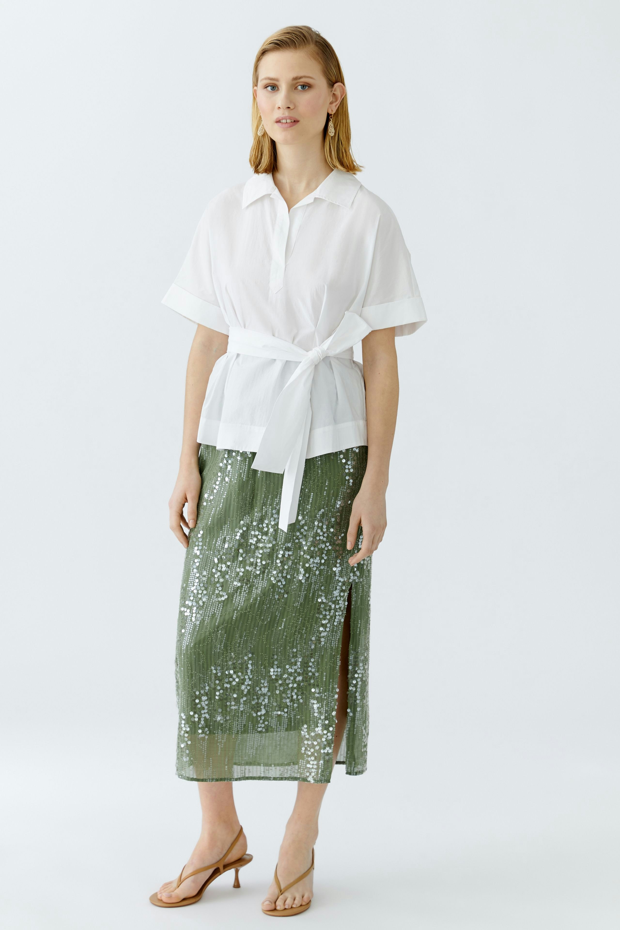 Moderne Damen-Bluse: Frau in weißer Bluse mit Gürtel und glitzerndem Rock steht lässig.