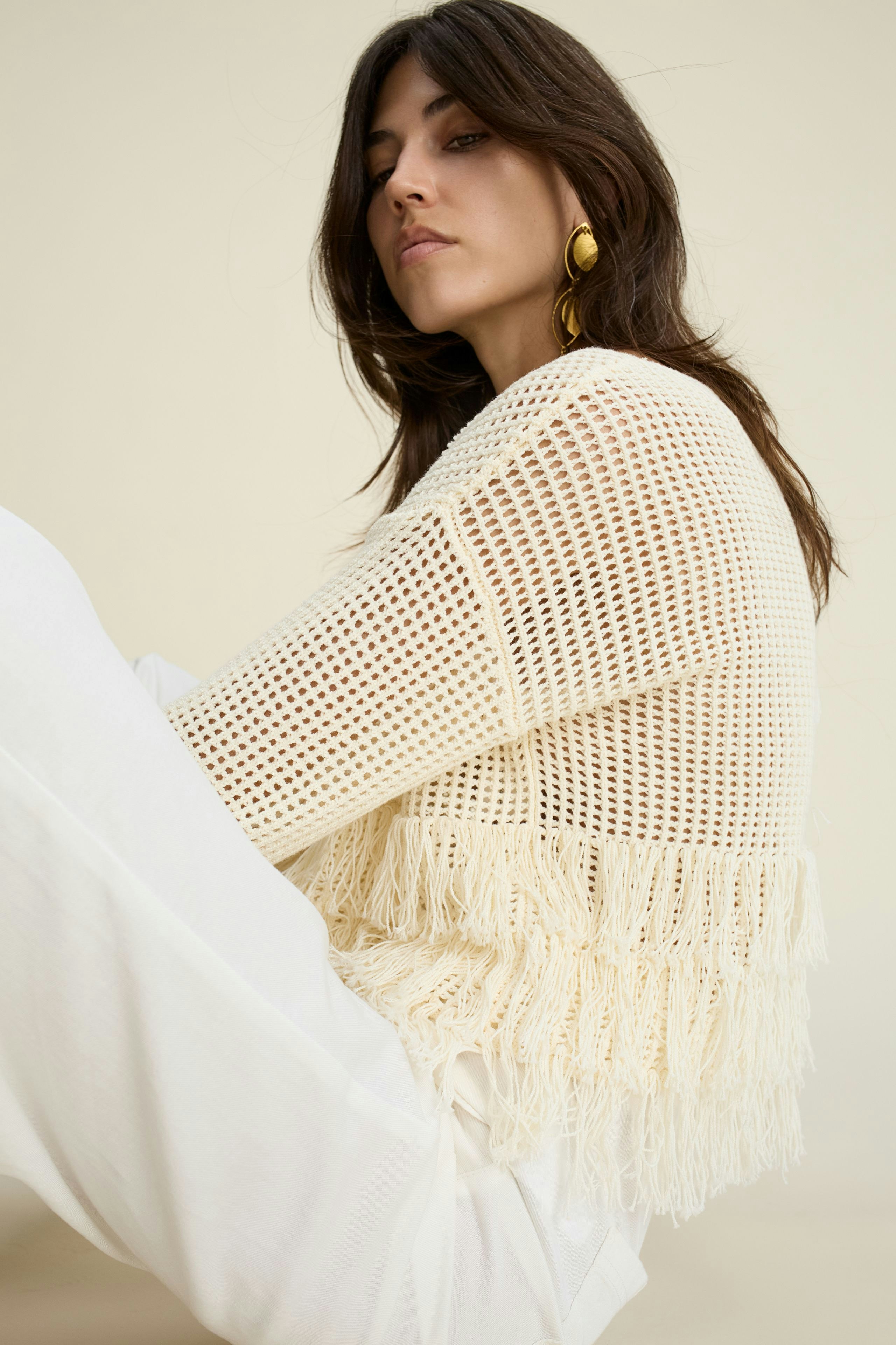 Damen-Pullover: Frau in einem hellen, luftigen Strickpullover mit Fransen sitzt entspannt.