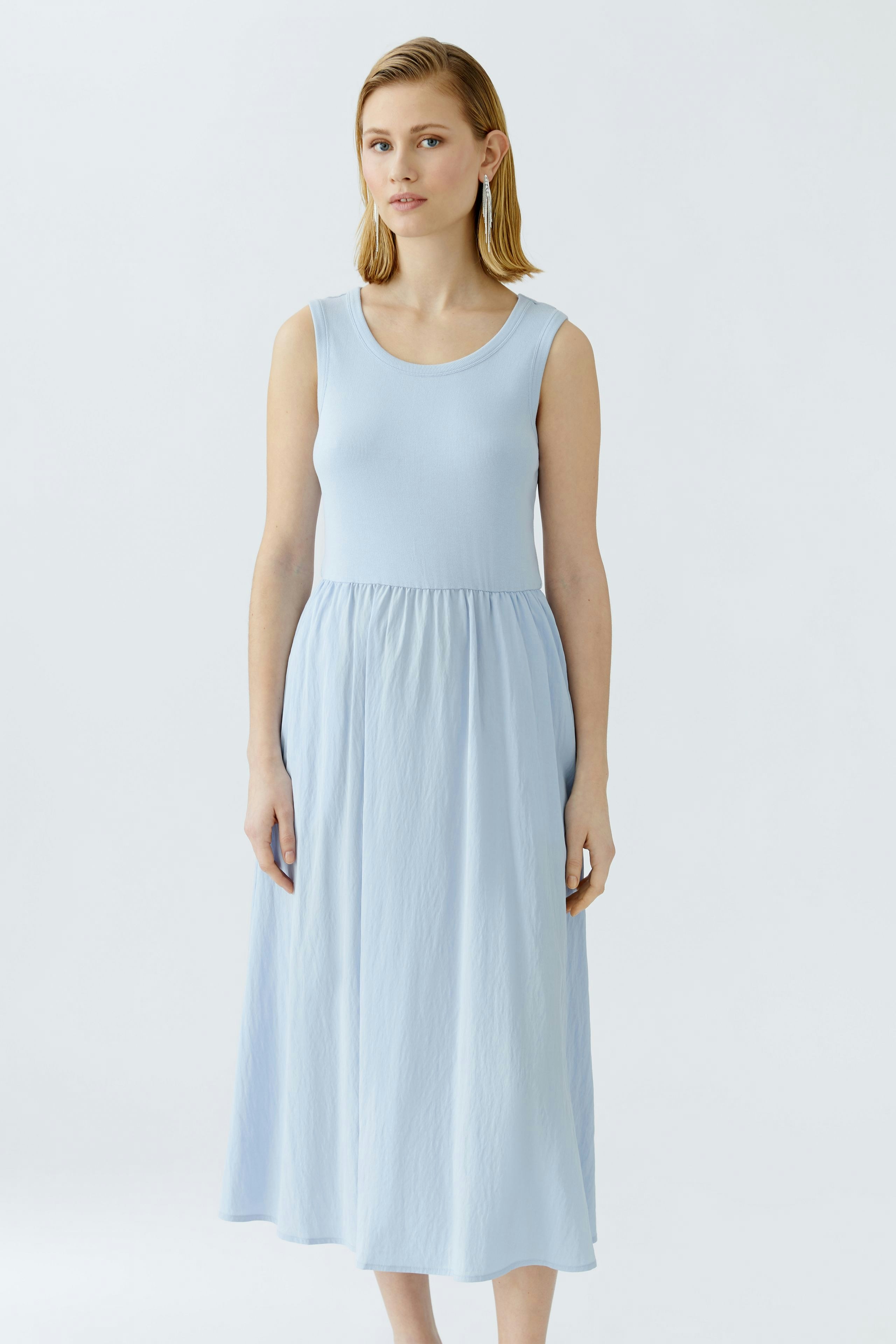 Luftiges Damen-Kleid: Frau in hellblauem, ärmellosem Kleid mit fließendem Rock steht entspannt.