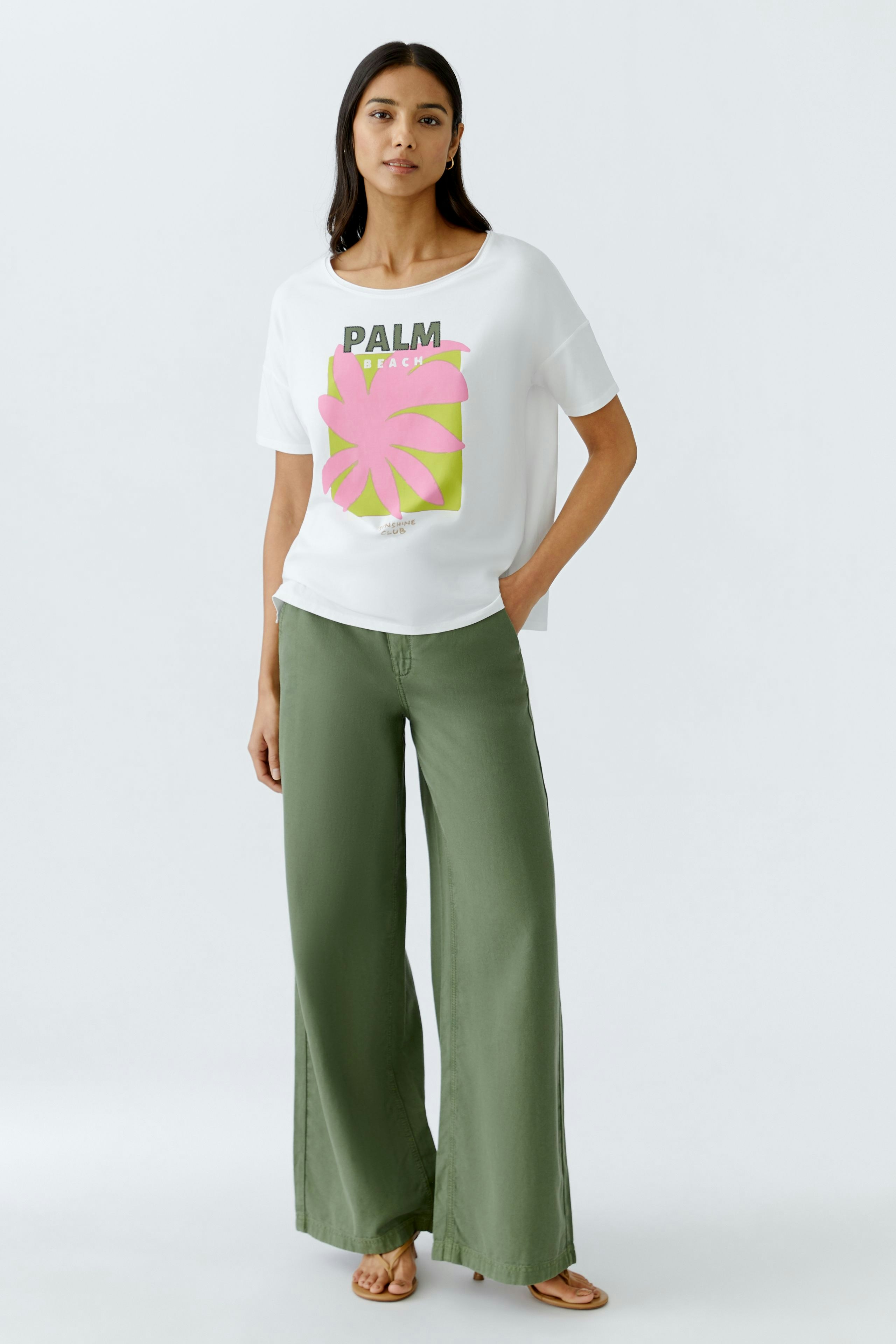 Stylish women's T-shirt: A woman in a white tee with a colorful palm print, paired with green wide-leg pants.