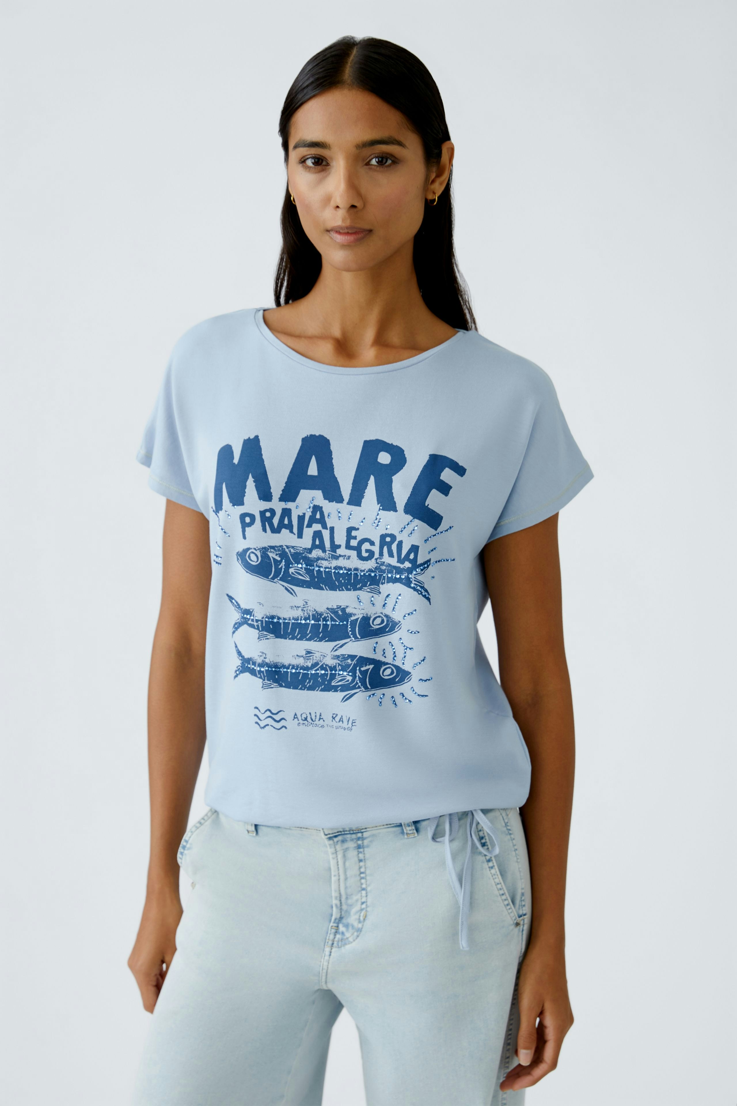 Casual women's T-shirt: Young woman in a light blue tee with a graphic print, paired with white pants.