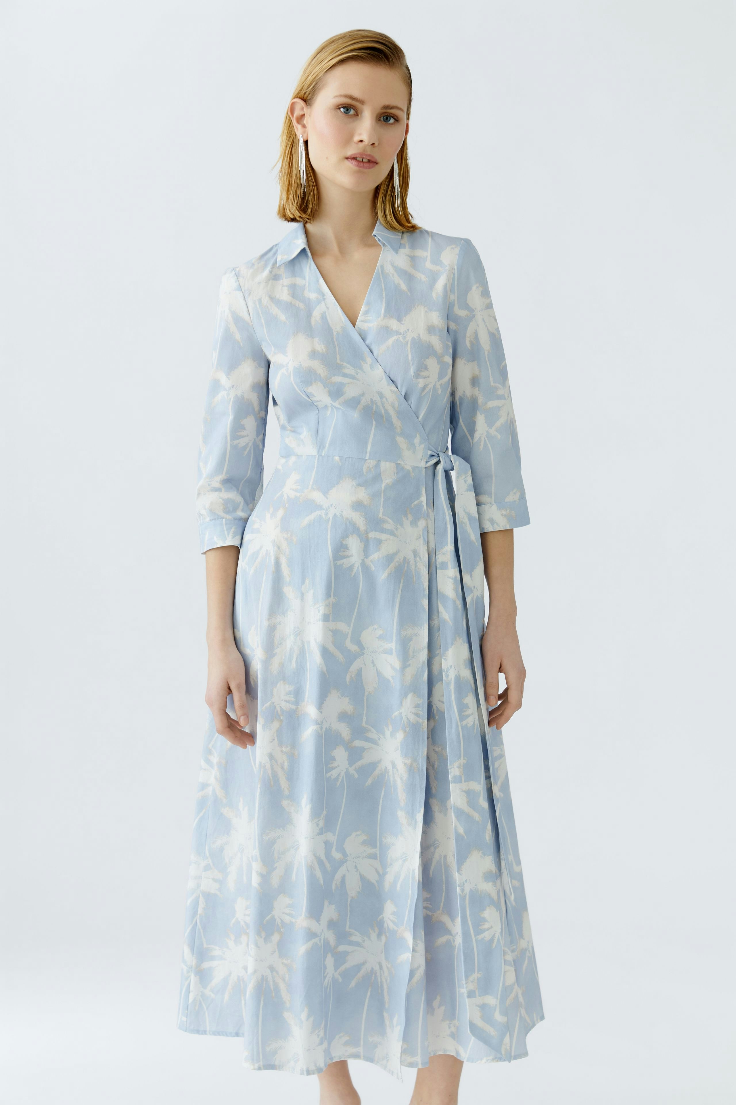 Wickelkleid: Frau in hellblauem Kleid mit floralem Muster und langen Ärmeln steht aufrecht.