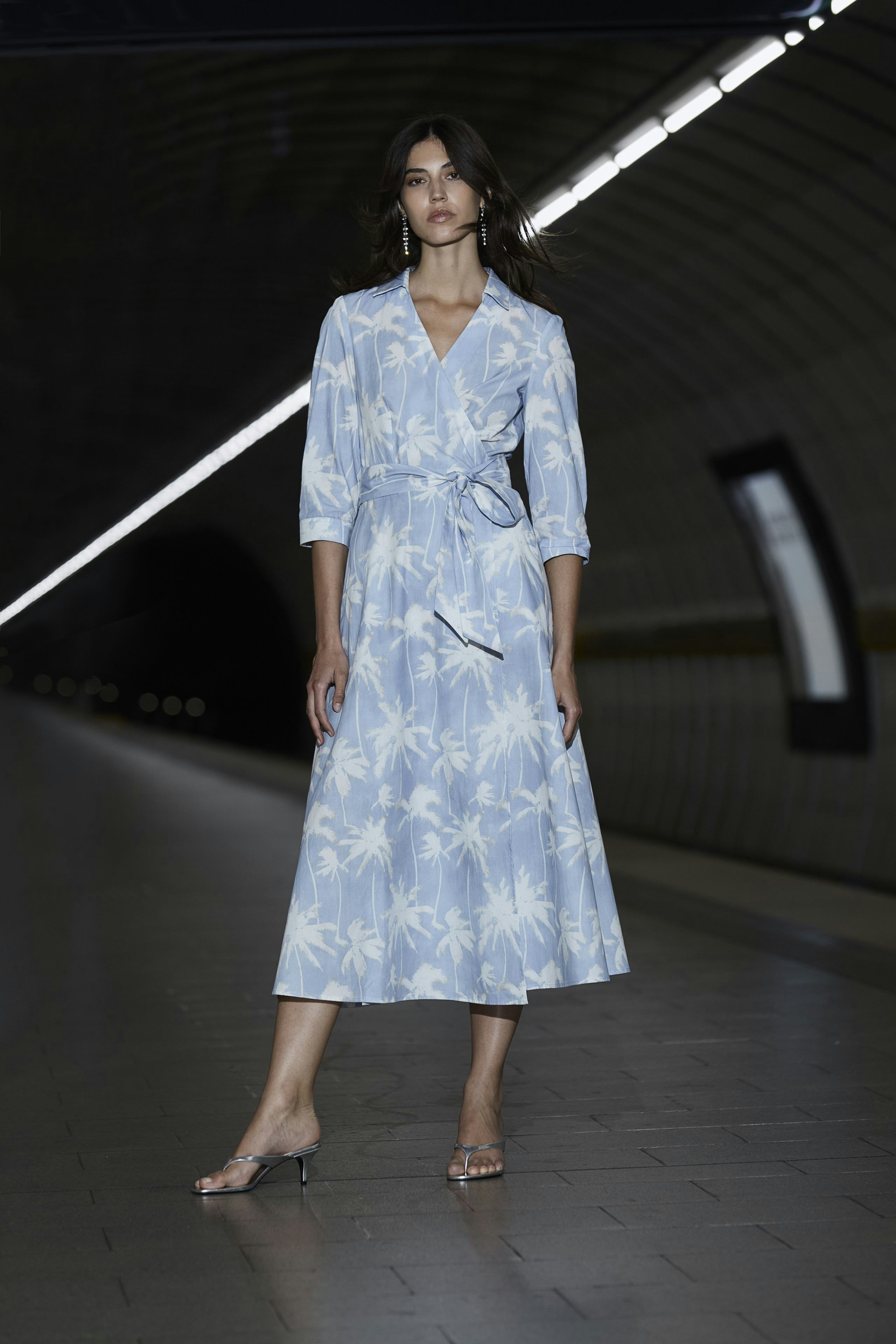 Wickelkleid: Frau in hellblauem Kleid mit floralem Muster, lässig und modern, steht in einem U-Bahn-Bereich.