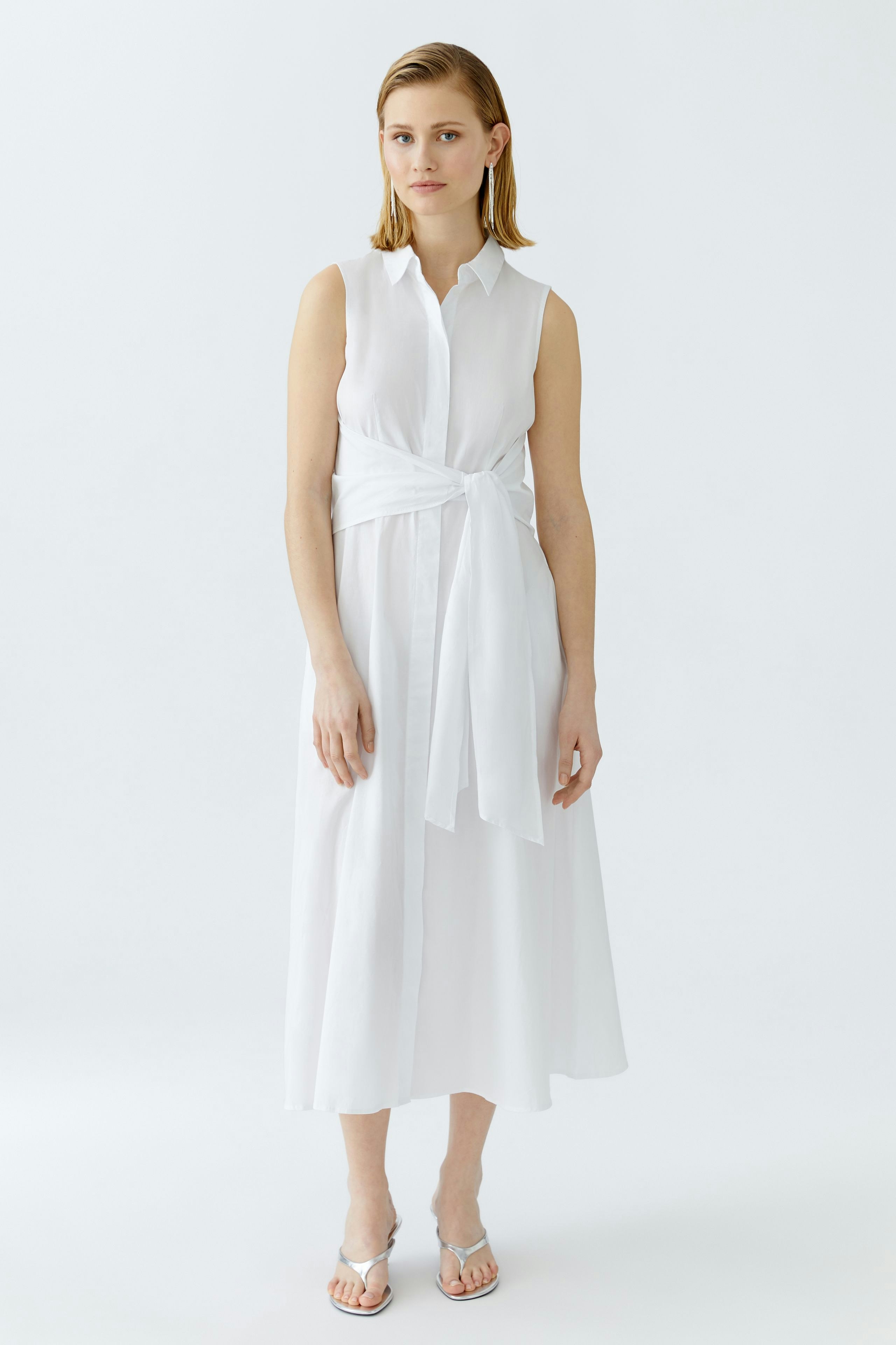 Maxikleid: Junge Frau in einem weißen, ärmellosen Maxikleid mit Taillengürtel steht aufrecht.