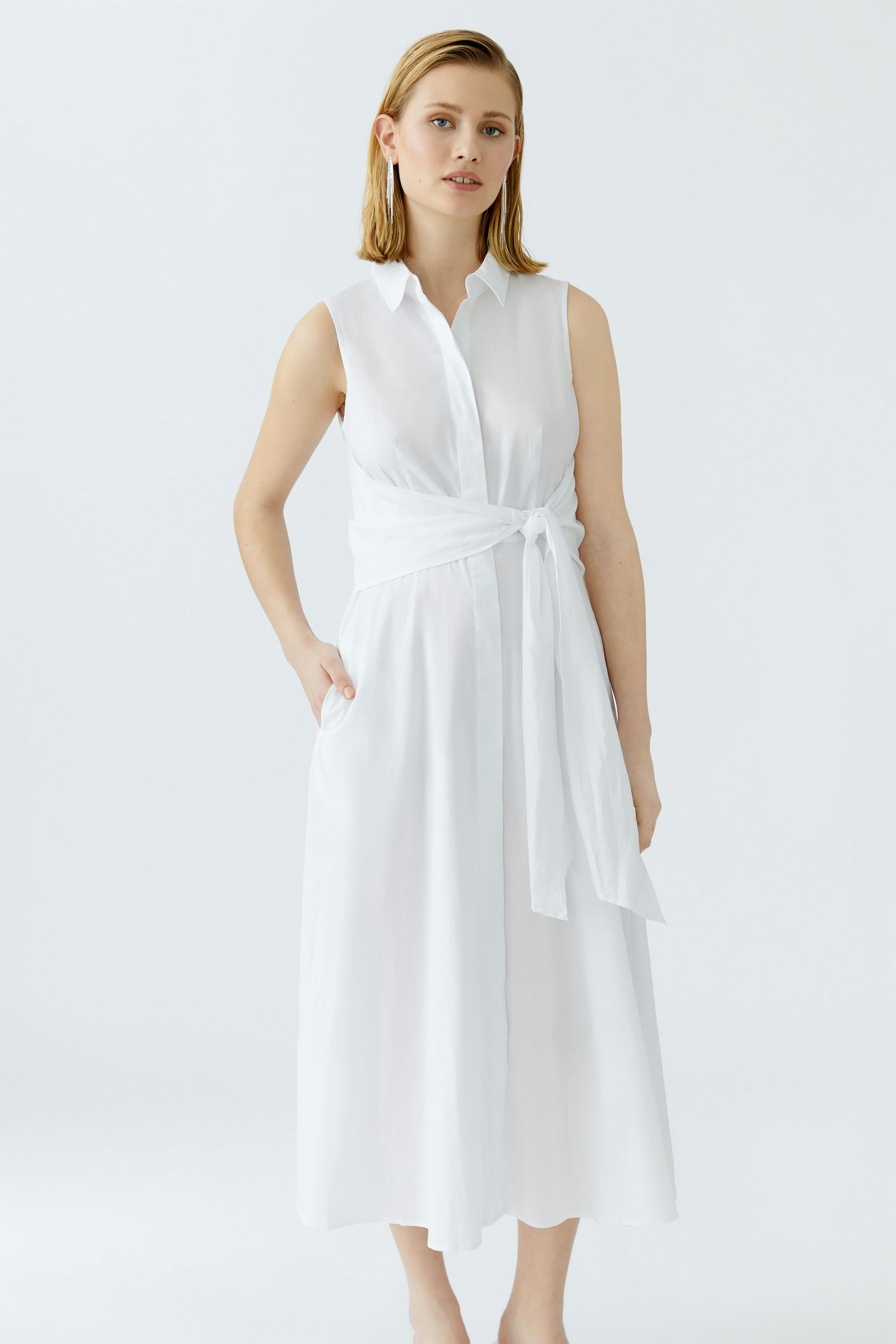 Maxikleid: Frau in einem weißen, ärmellosen Kleid mit Gürtel und Hemdkragen, lässig und modern.