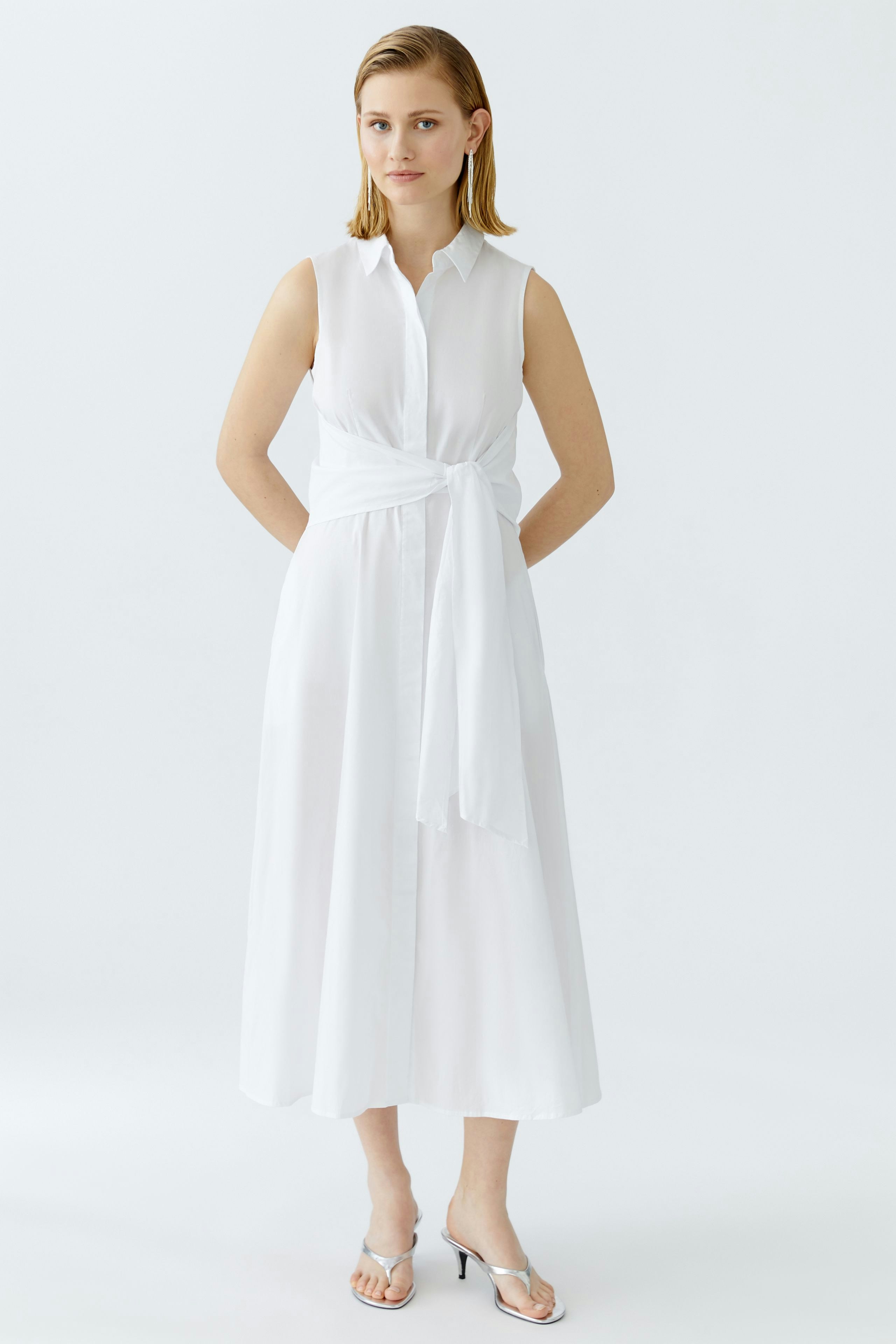 Maxikleid: Junge Frau in einem weißen, ärmellosen Kleid mit Taillengürtel steht aufrecht.