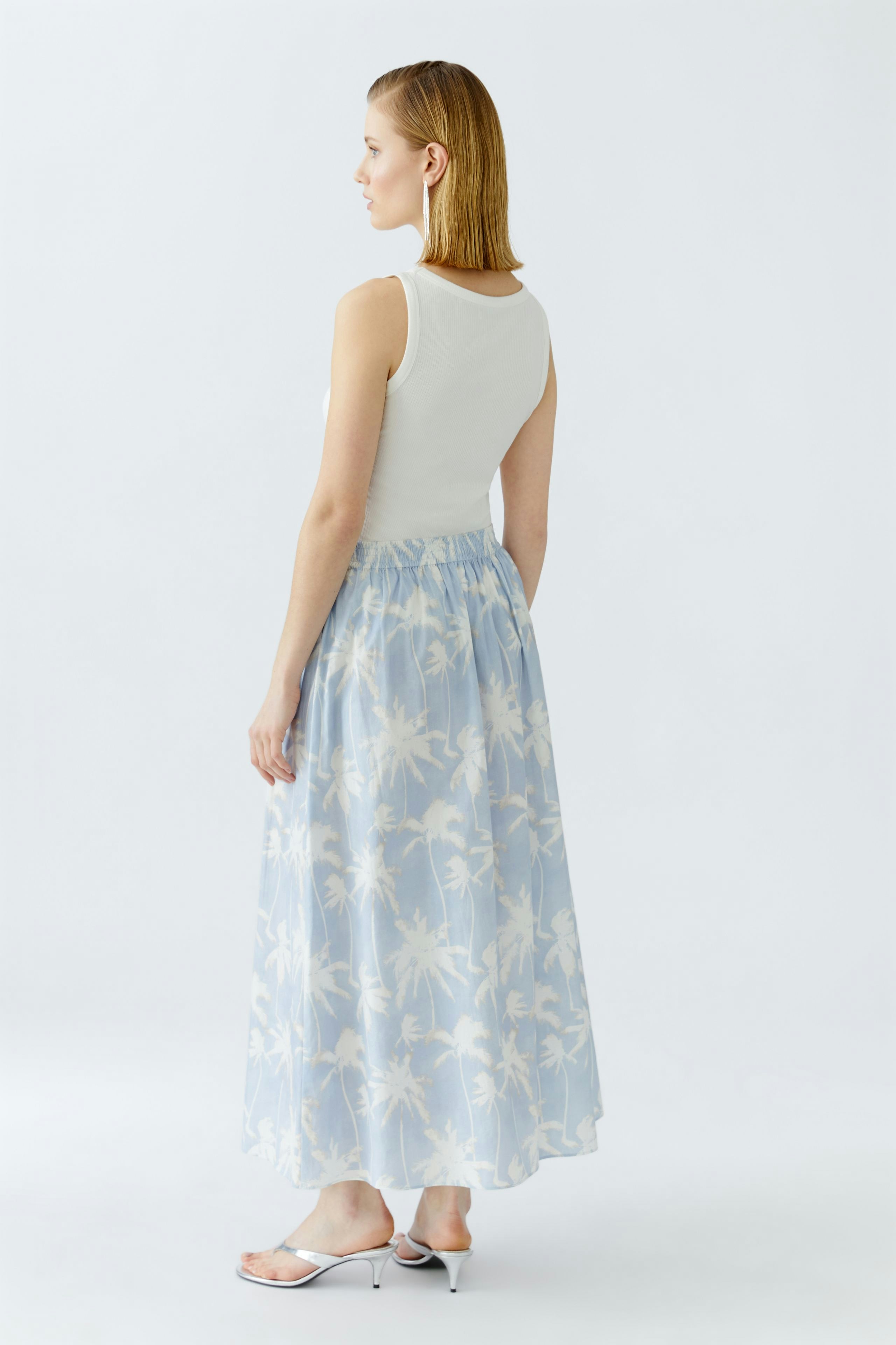 Light blue maxi skirt with palm tree print, worn by a woman, featuring a comfortable elastic waistband.