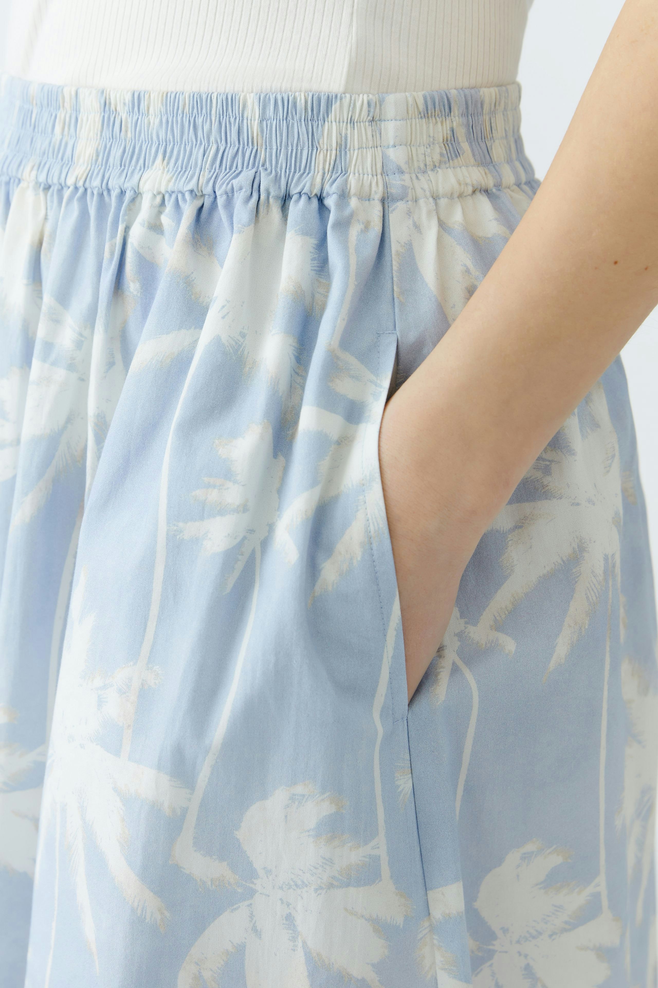 Light blue maxi skirt with palm tree print and pockets, worn by a woman.