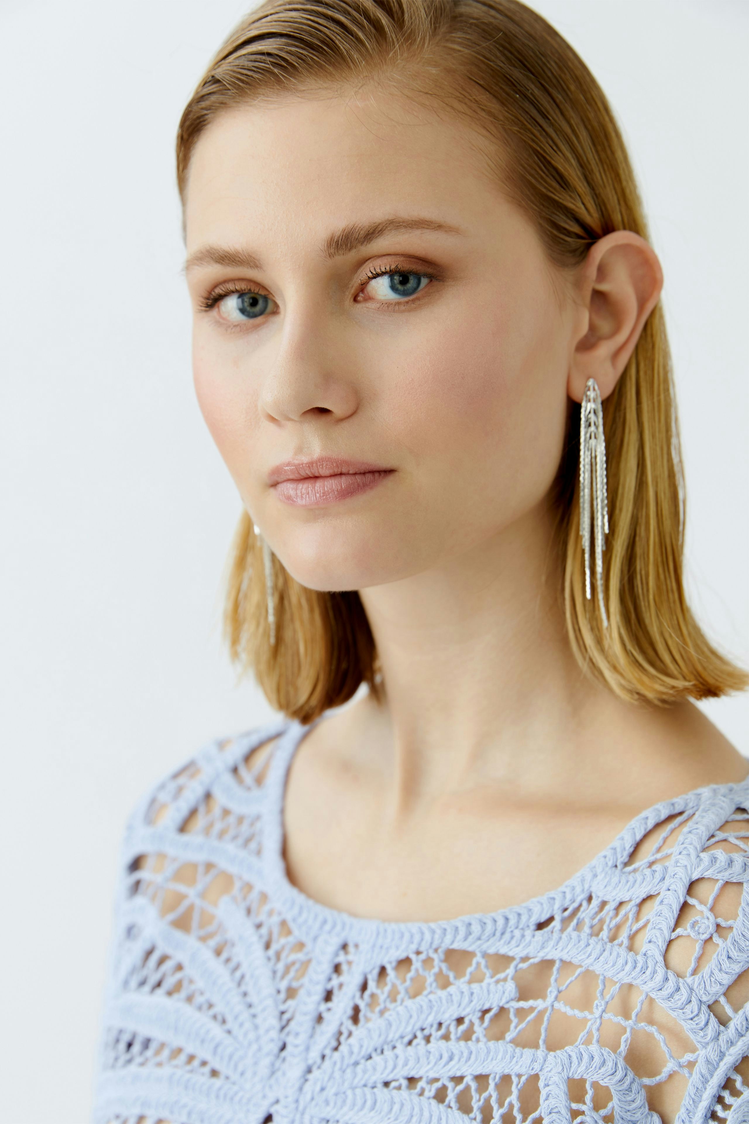 Damen-Pullover: Junge Frau mit hellblauem, gehäkeltem Pullover und langen Ohrringen blickt freundlich.
