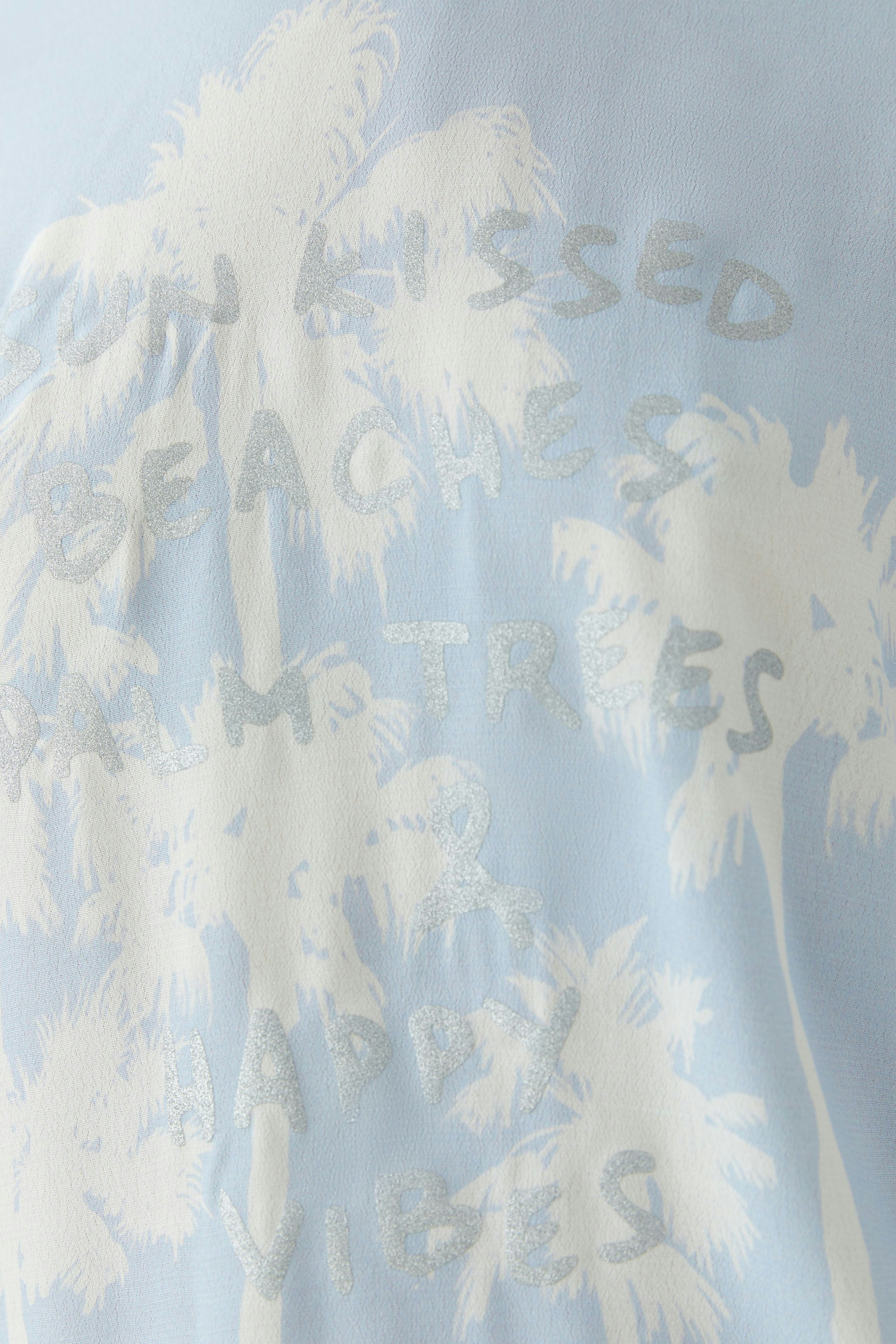 Luftiges Blusenshirt für Damen in sanftem Blau mit Palmen-Print und lässigem Schriftzug.