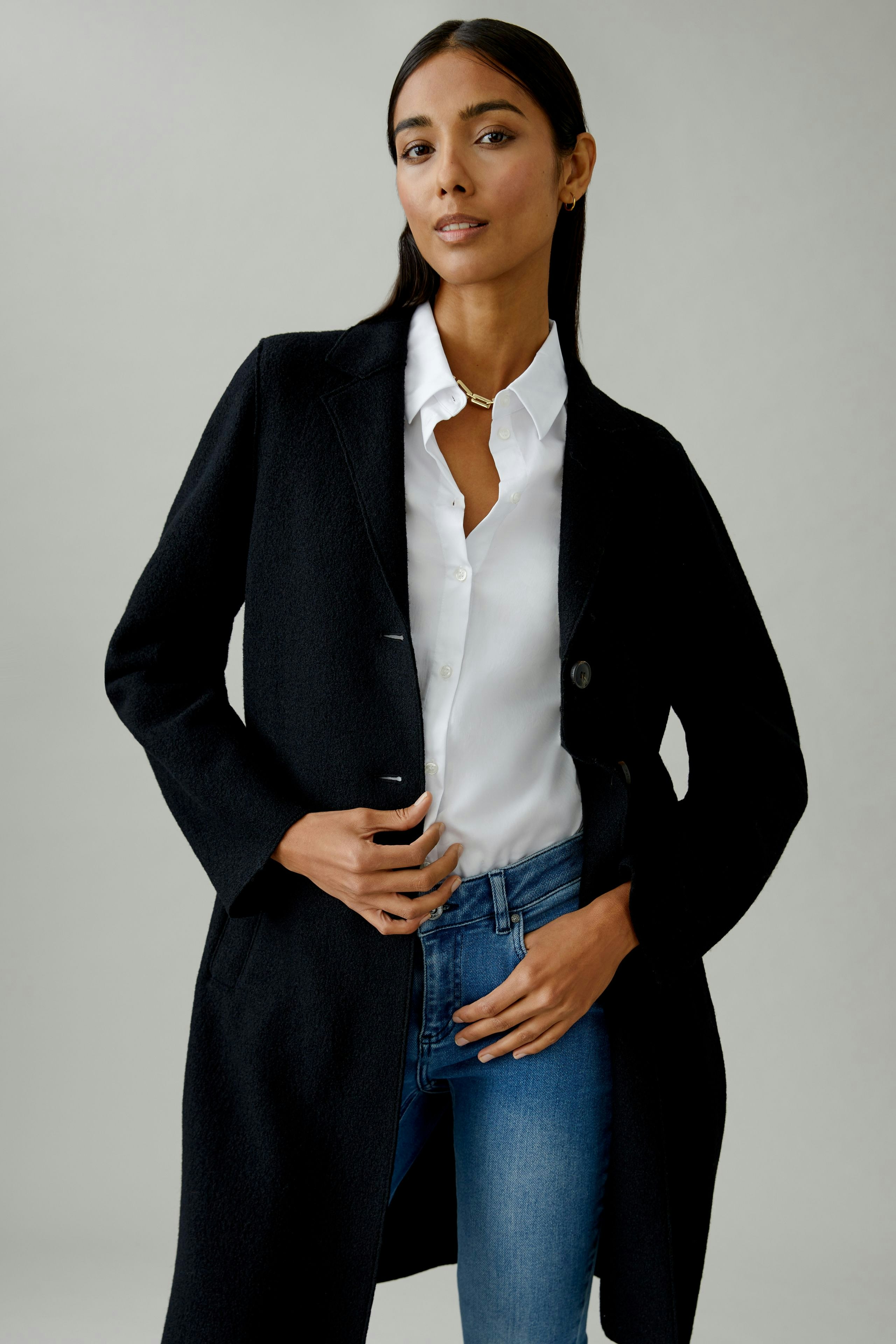 Damen Wollmantel: Frau in schwarzem Wollmantel mit weißem Hemd und Jeans, lässig und modern.