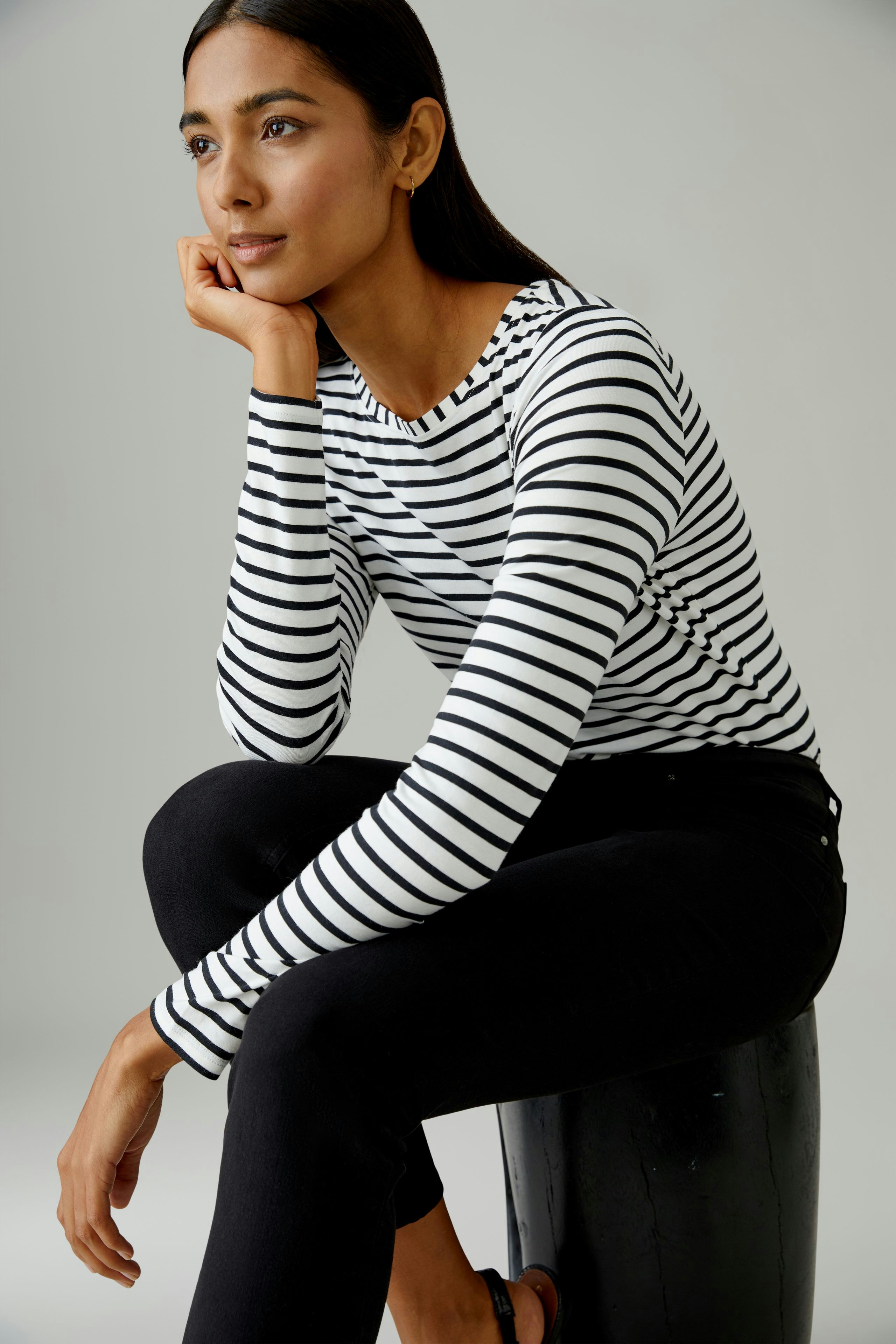 Langarmshirt: Junge Frau in gestreiftem Langarmshirt sitzt nachdenklich auf einem Hocker.