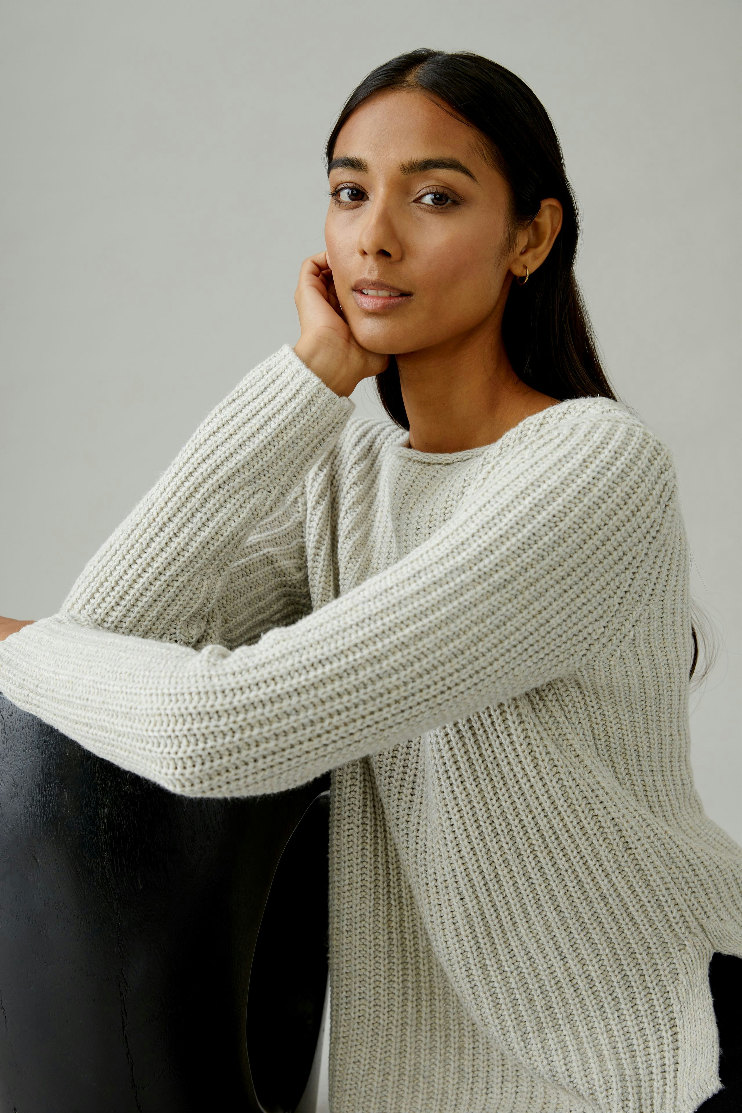Casual women's pullover: Young woman in a light gray knitted pullover, resting her chin on her hand.