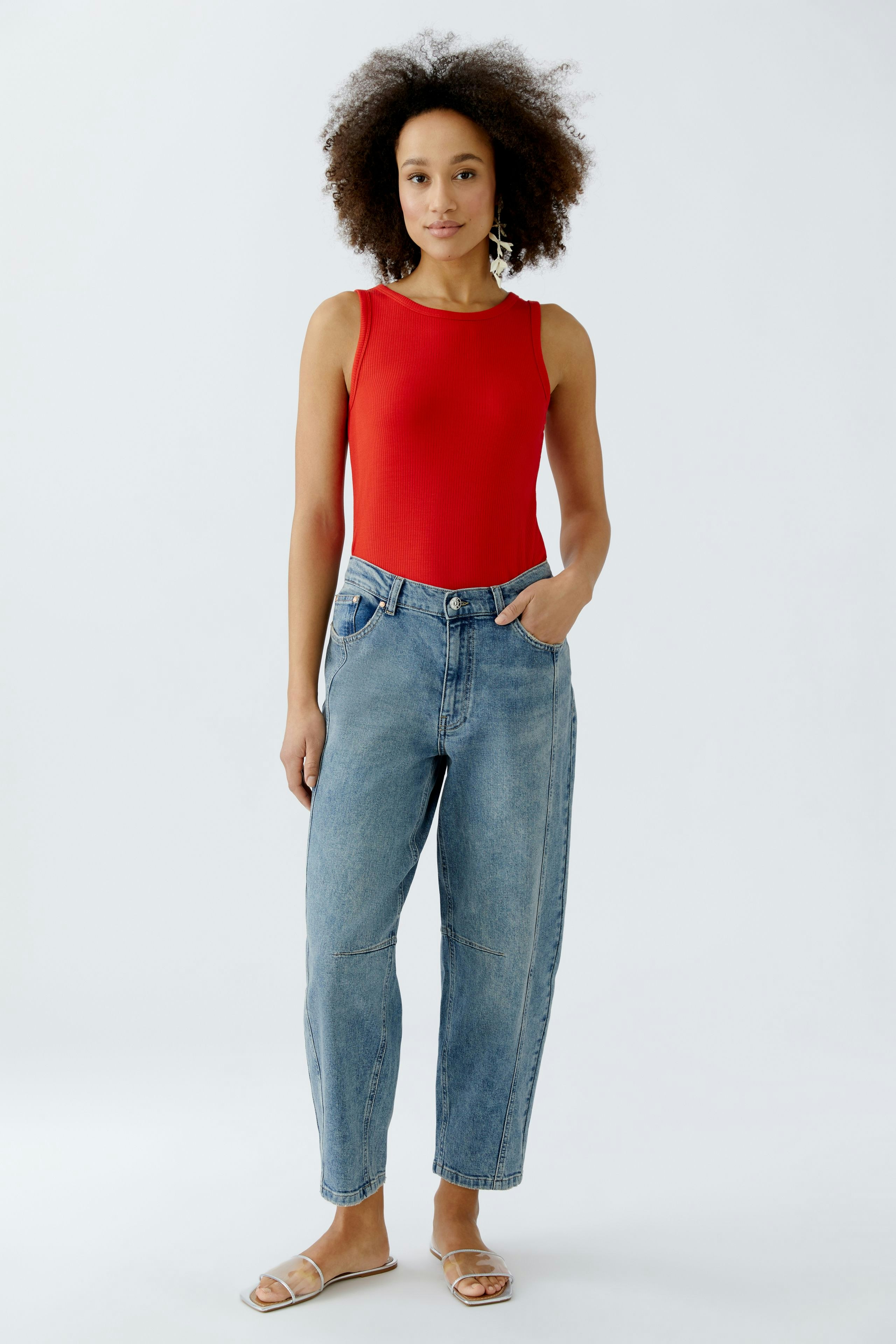 Casual women's top: Young woman in a red sleeveless top and relaxed-fit jeans stands confidently.