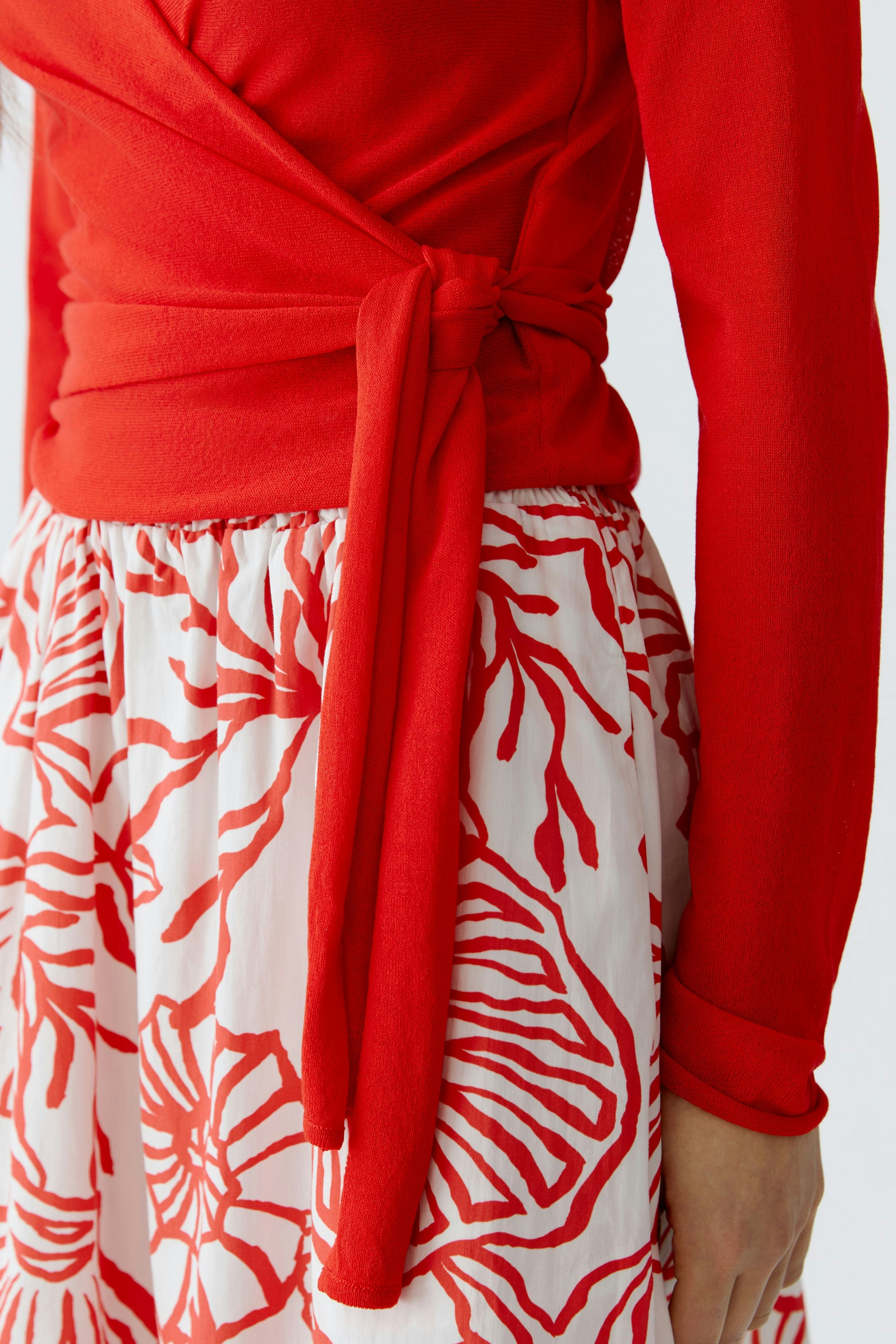 Damen-Strickjacke: Frau in roter Strickjacke mit Bindegürtel, kombiniert mit einem blumigen Rock.