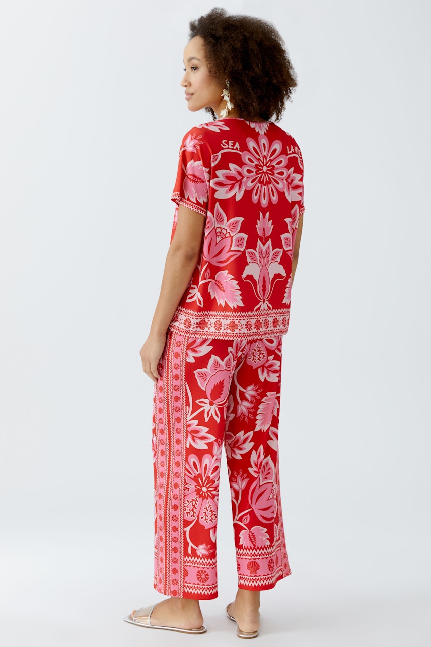 Damen-T-Shirt: Frau in rotem T-Shirt mit floralem Muster und lässiger Hose, von hinten zu sehen.