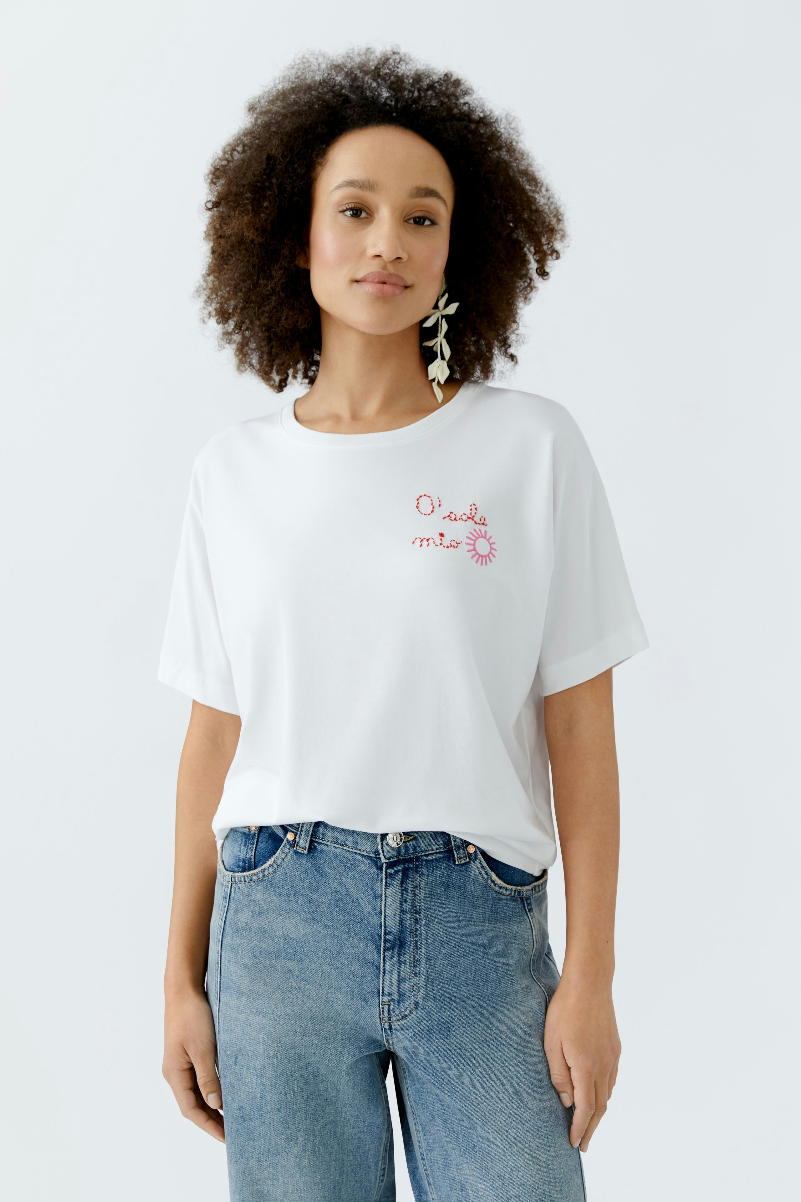Weißes Damen-T-Shirt mit buntem Aufdruck, lässig kombiniert mit Jeans und großen Ohrringen.