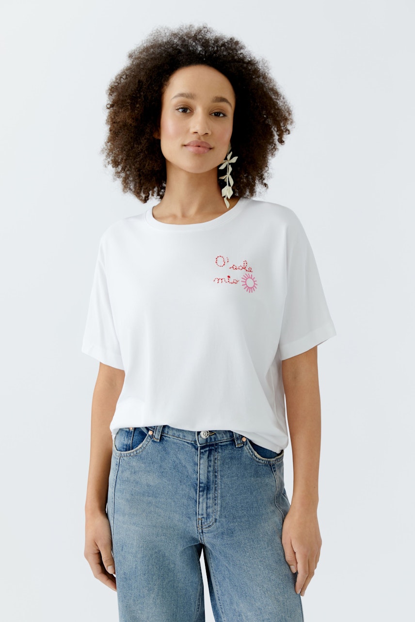 Weißes Damen-T-Shirt mit buntem Aufdruck, lässig kombiniert mit Jeans und großen Ohrringen.