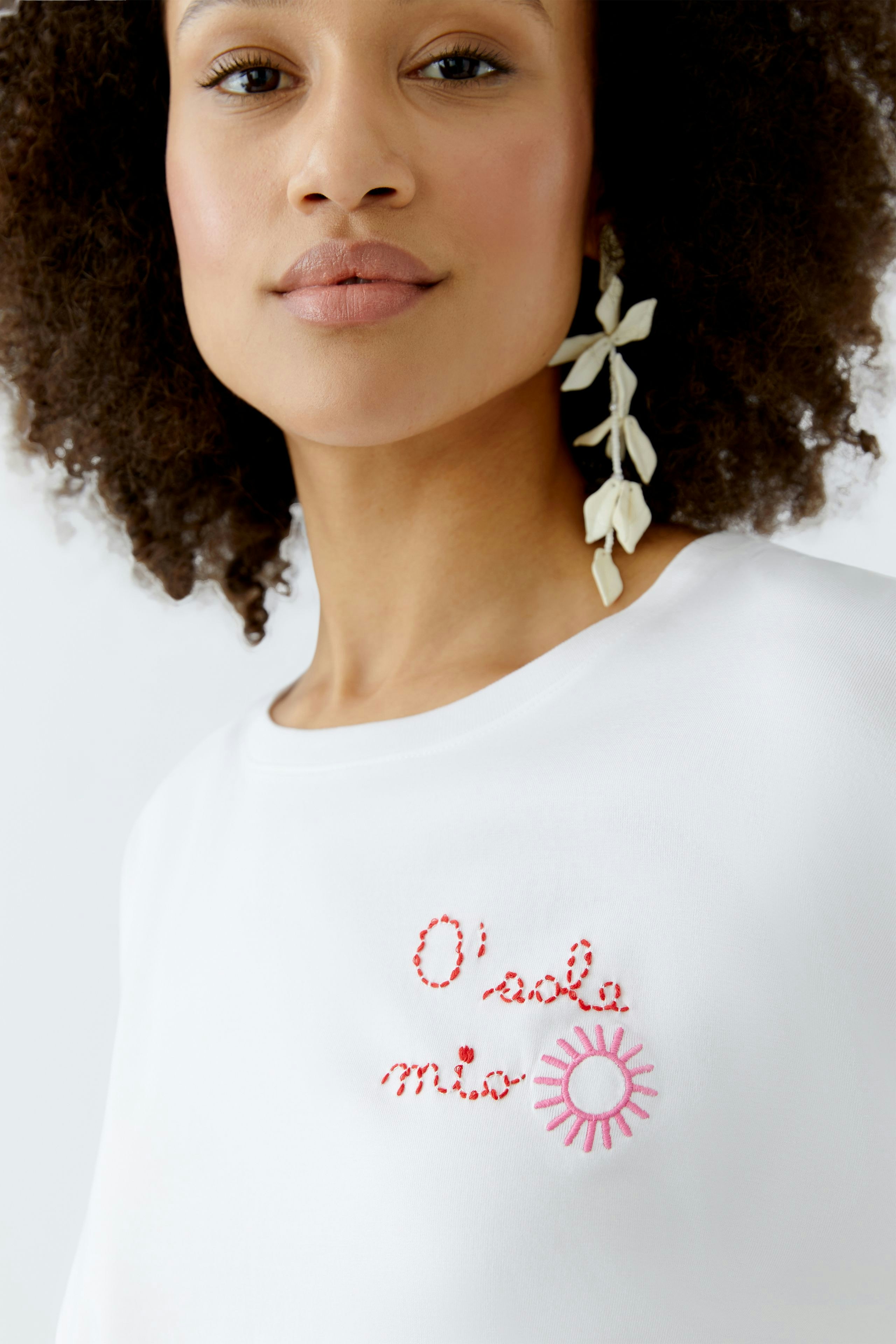 Damen-T-Shirt: Frau trägt ein weißes Shirt mit buntem, gesticktem Schriftzug und Sonnenmotiv.