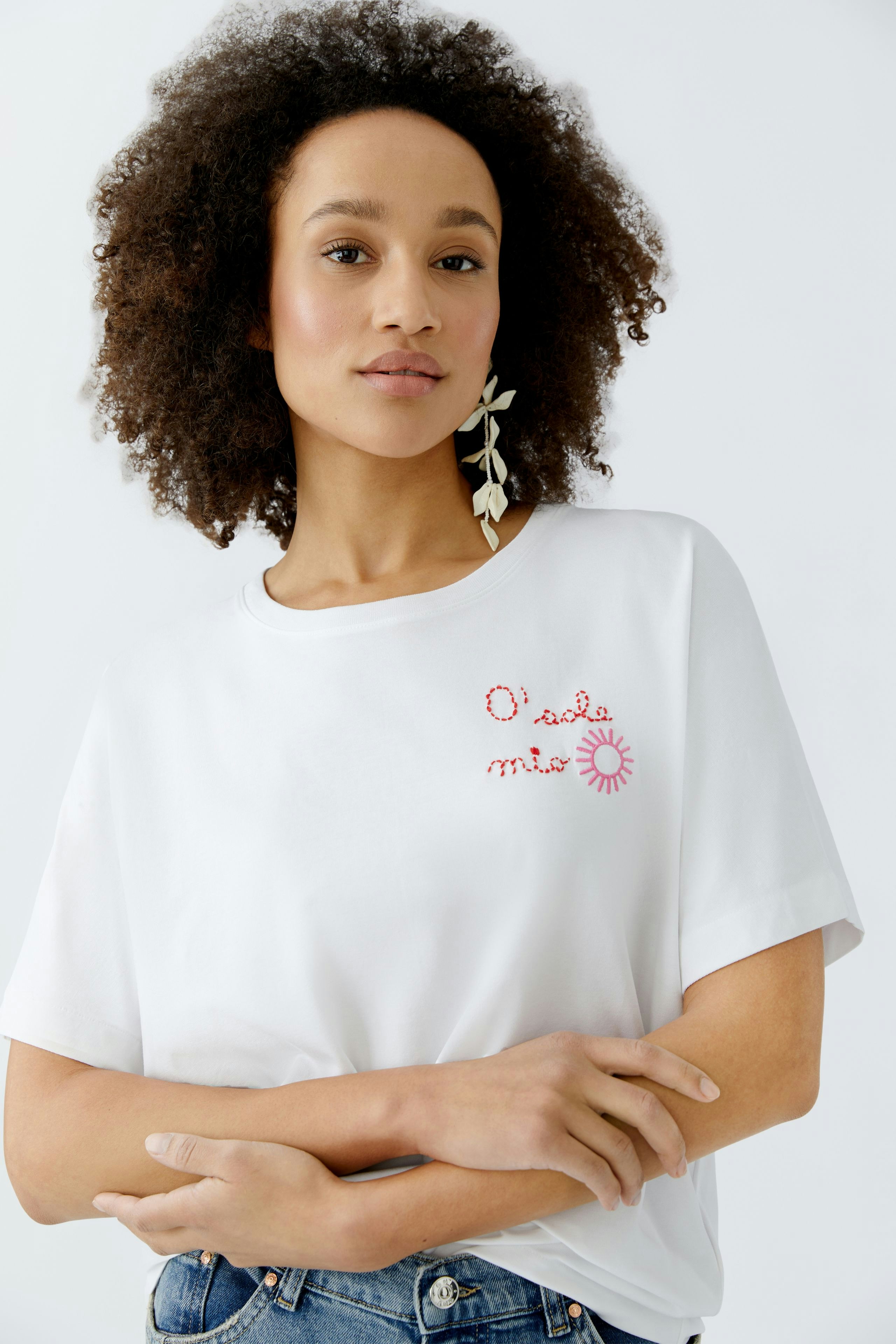 Damen-T-Shirt: Frau mit lockigem Haar trägt ein weißes T-Shirt mit buntem Schriftzug und lässigen Jeans.