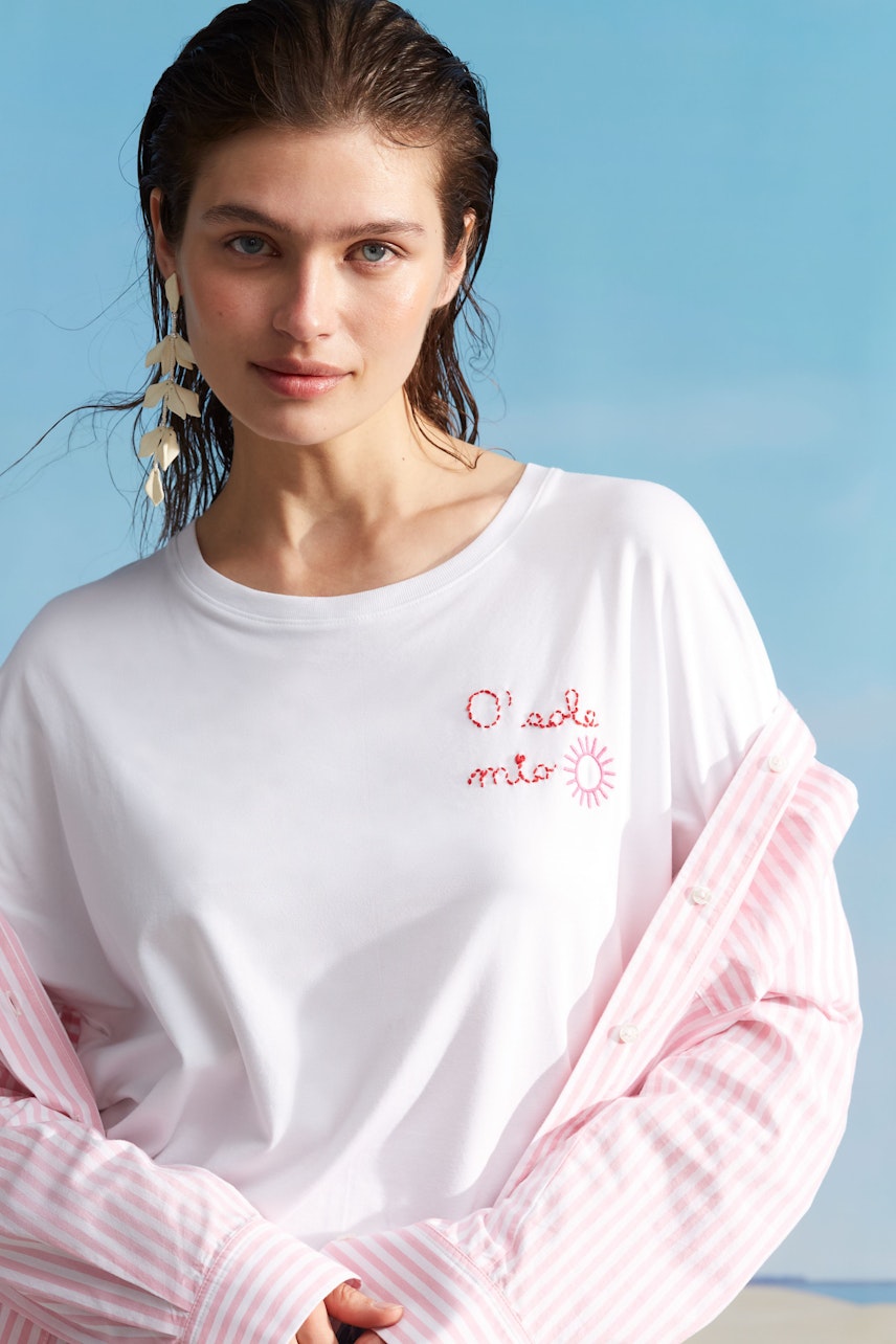 Damen-T-Shirt: Junge Frau trägt ein weißes T-Shirt mit roter Stickerei und lässiger gestreifter Bluse.
