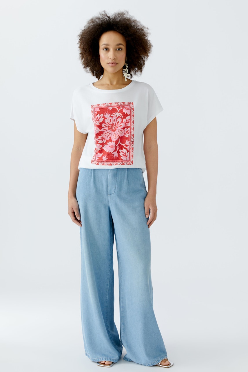 Damen-T-Shirt: Frau trägt ein weißes T-Shirt mit rotem Blumenmotiv und lässigen Jeans.