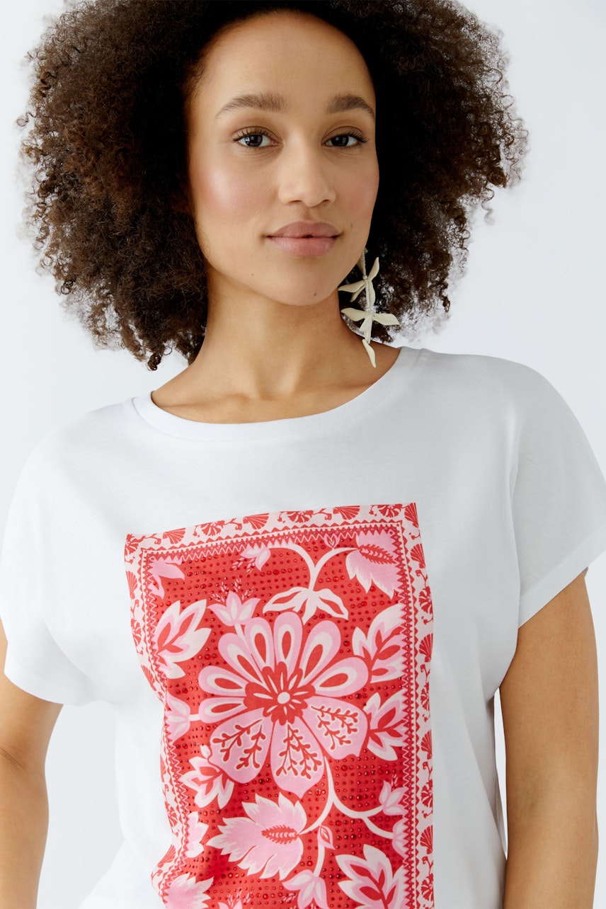 Damen-T-Shirt: Frau trägt ein weißes Shirt mit rotem Blumenmuster und lässigem Schnitt.