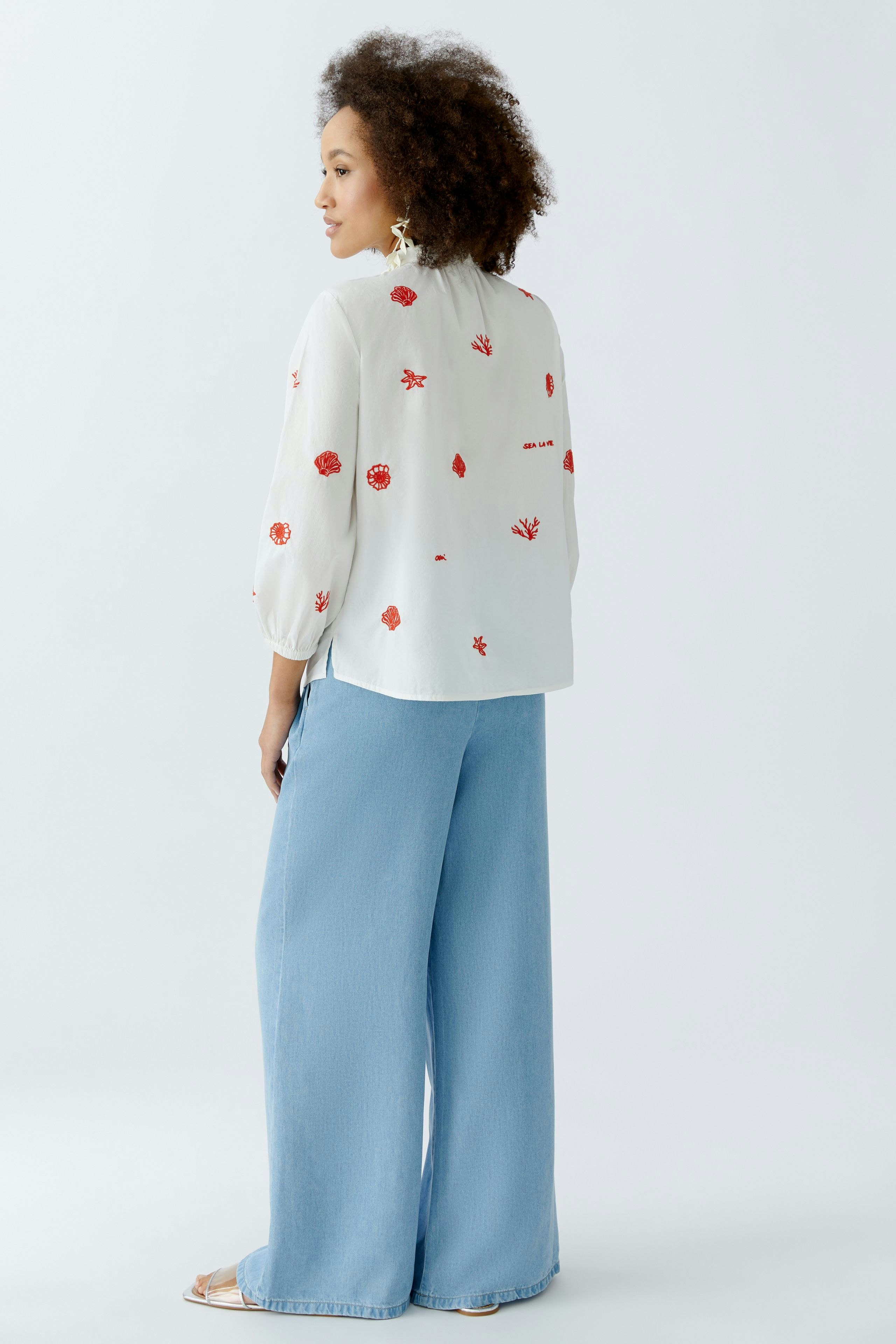 Damen-Bluse: Frau mit lockigen Haaren trägt eine weiße Bluse mit roten Blumenstickereien und lässigen Jeans.