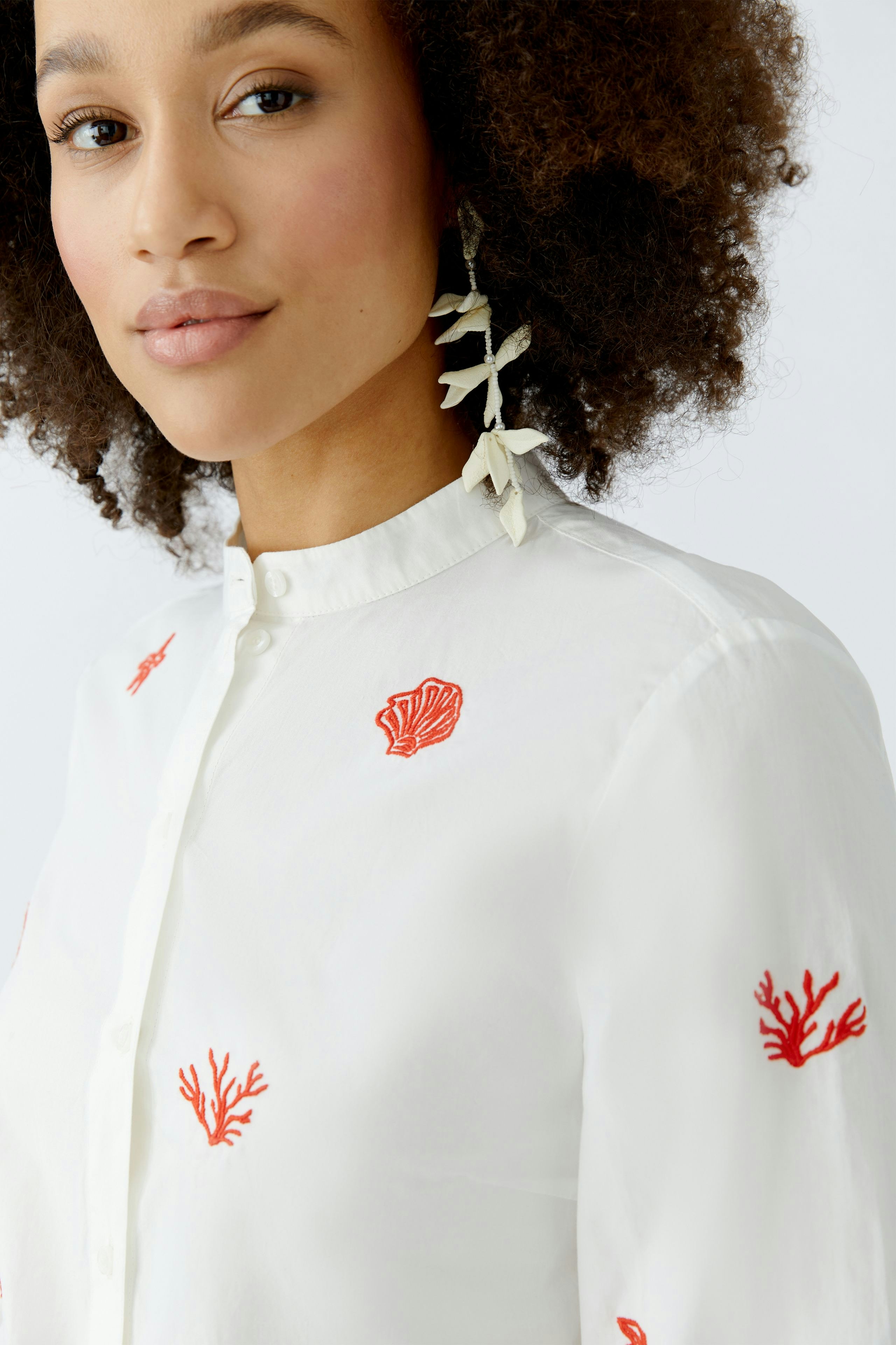Damen-Bluse: Frau mit lockigen Haaren trägt eine weiße Bluse mit roten Korallenstickereien.