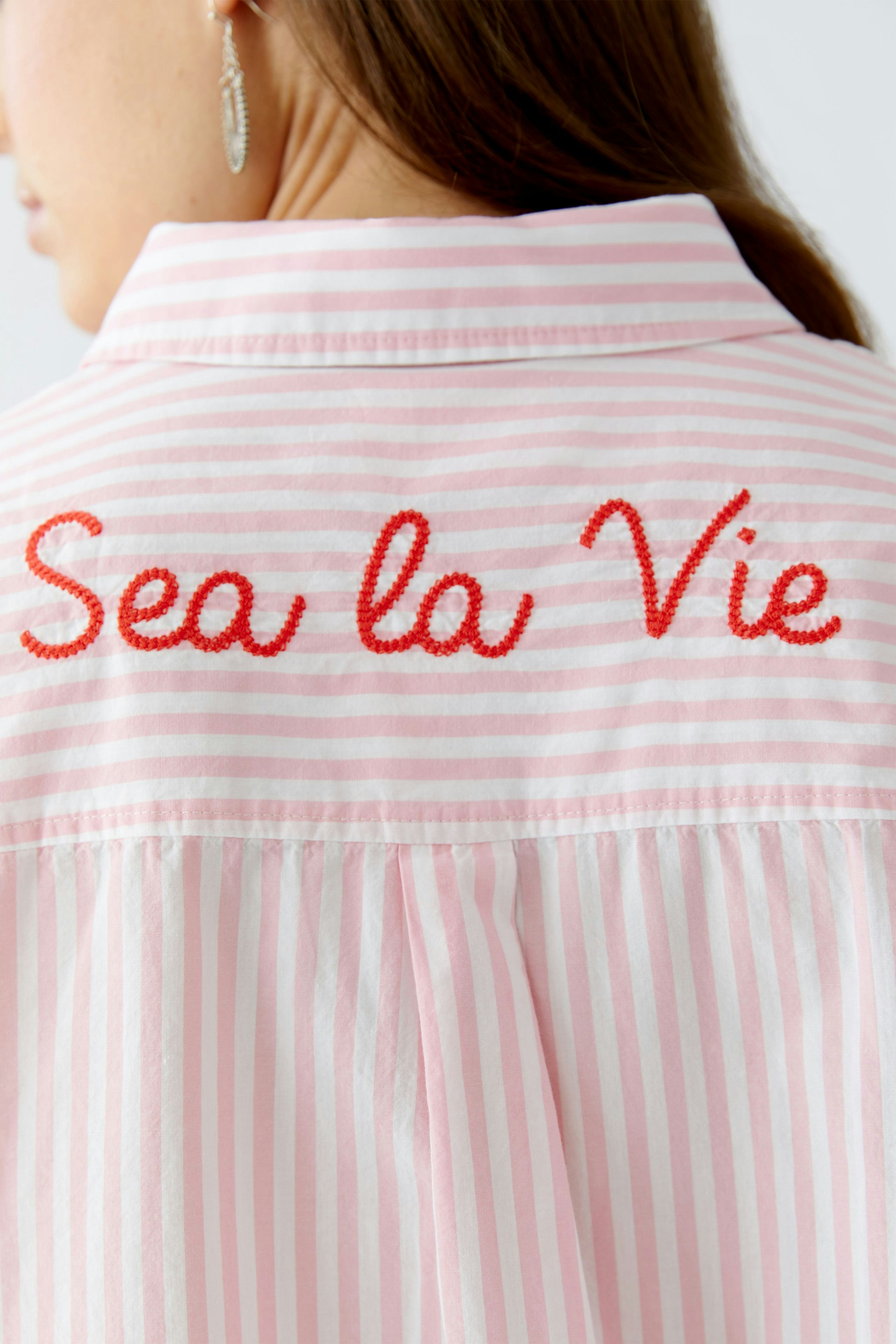 Hemdbluse: Frau trägt eine gestreifte Bluse mit roter Stickerei "Sea la Vie".